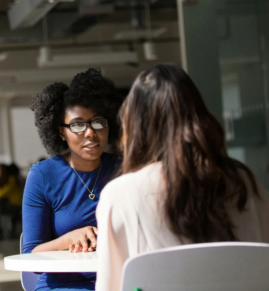 Two women sitting at a table engaged in conversation, one facing the camera and wearing glasses and a blue top, the other with back to camera and brown hair, in an indoor setting.