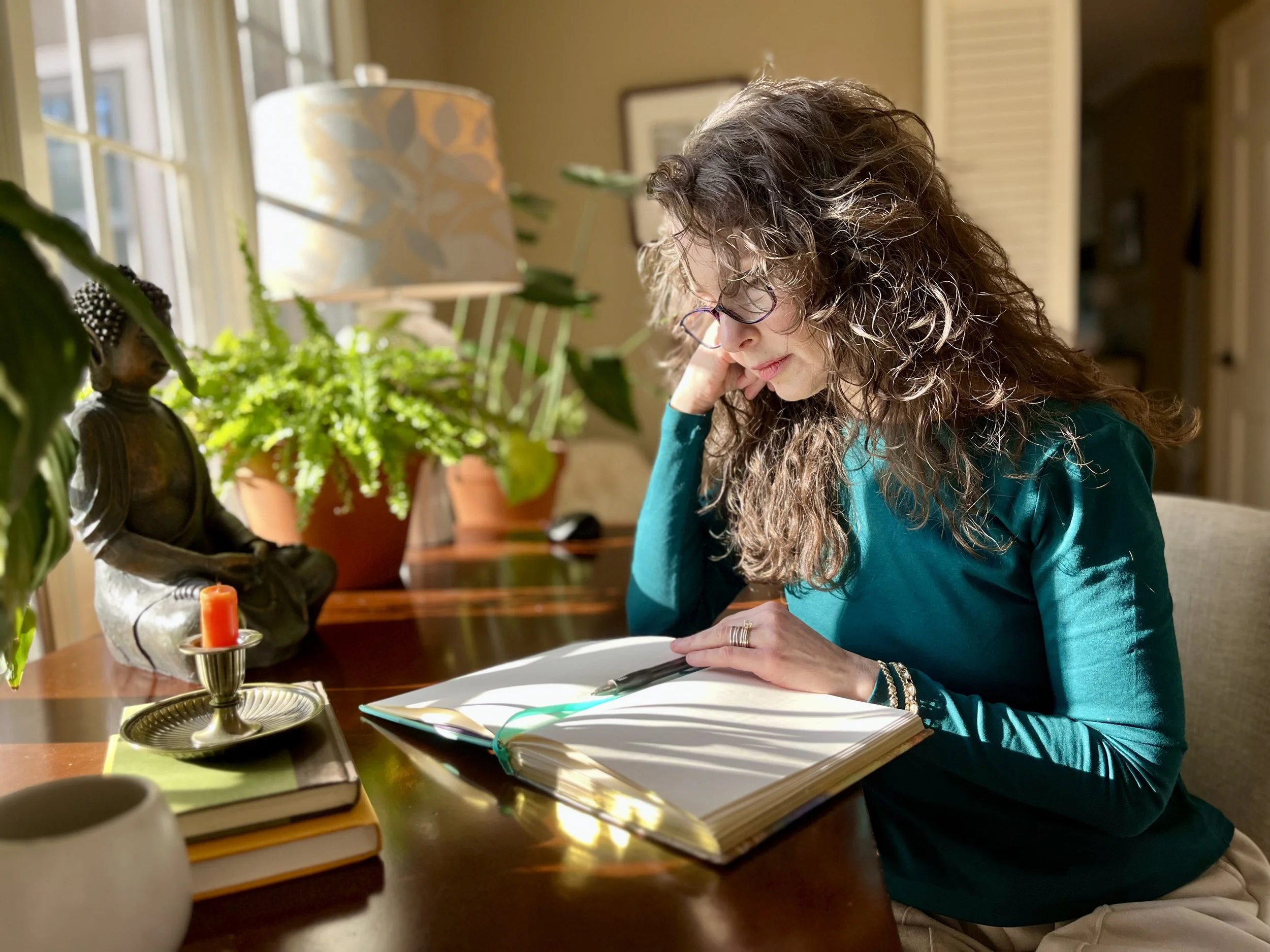 Woman with curly hair and glasses sitting at a desk, reading a notebook. The desk is decorated with a small Buddha statue, a candle, a stack of books, and a potted plant. Sunlight streams through a nearby window, casting light on the scene.