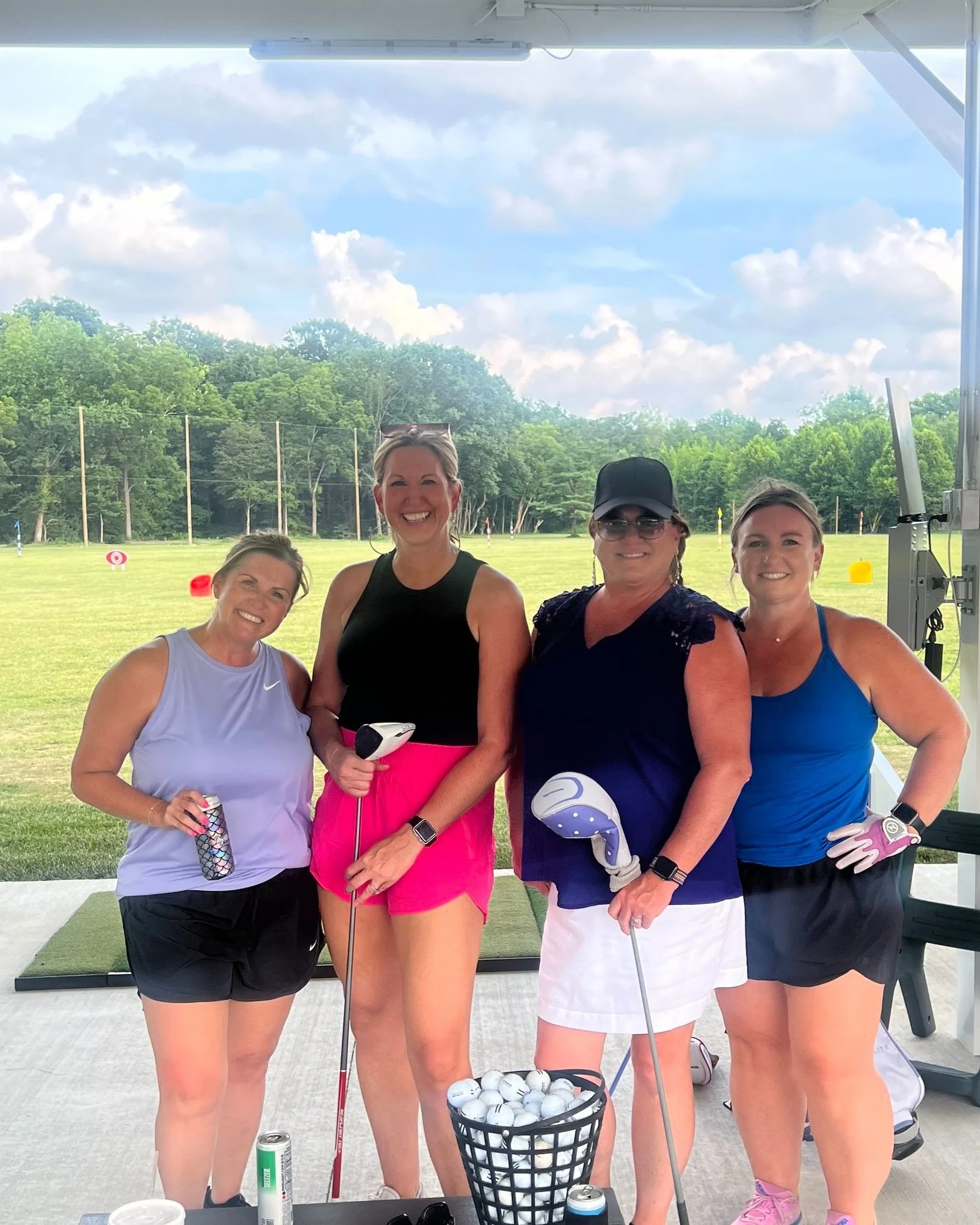 Four women standing together at a golf driving range, holding golf clubs and smiling. They are dressed in athletic wear, with a basket of golf balls in front of them and a scenic green golf course with trees in the background.