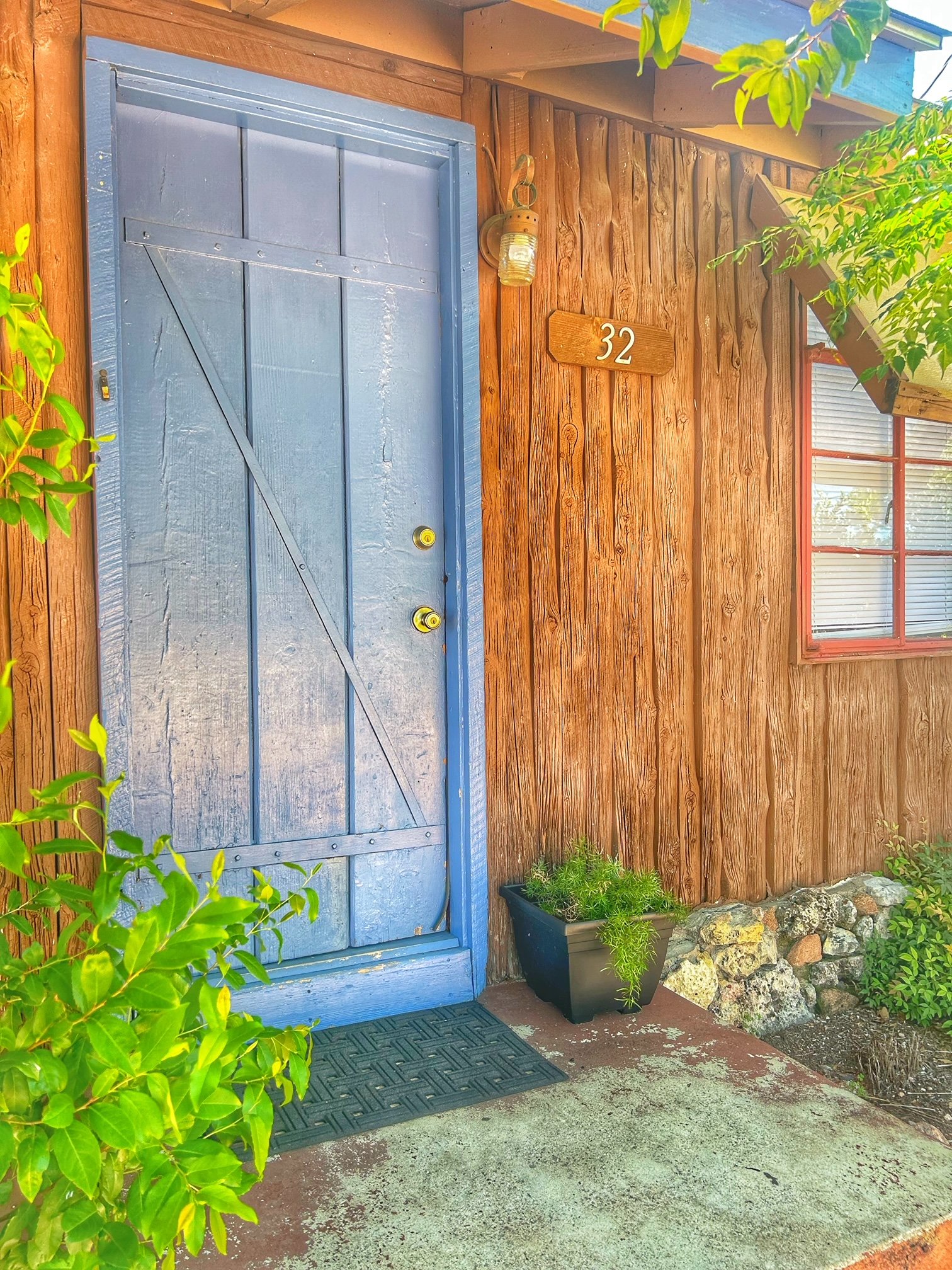 A small blue house with wooden accents, featuring a blue door and several windows. It has a covered porch with a corrugated metal roof supported by wooden beams. Stone landscaping and a small garden with flowers are situated near the entrance. A tree is visible in the background.