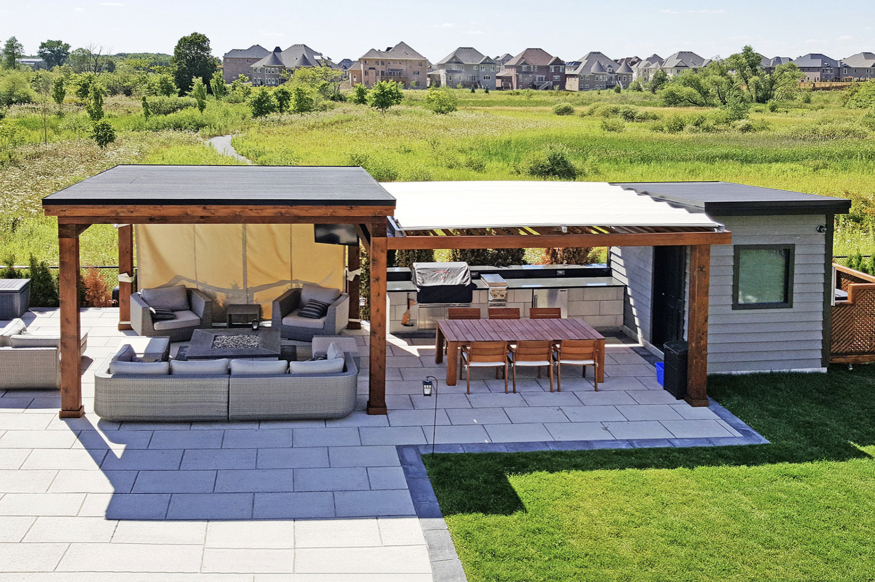 Outdoor patio with lounge seating, dining table, and barbecue area under a wooden pergola, surrounded by grass and a view of nearby houses and open field.