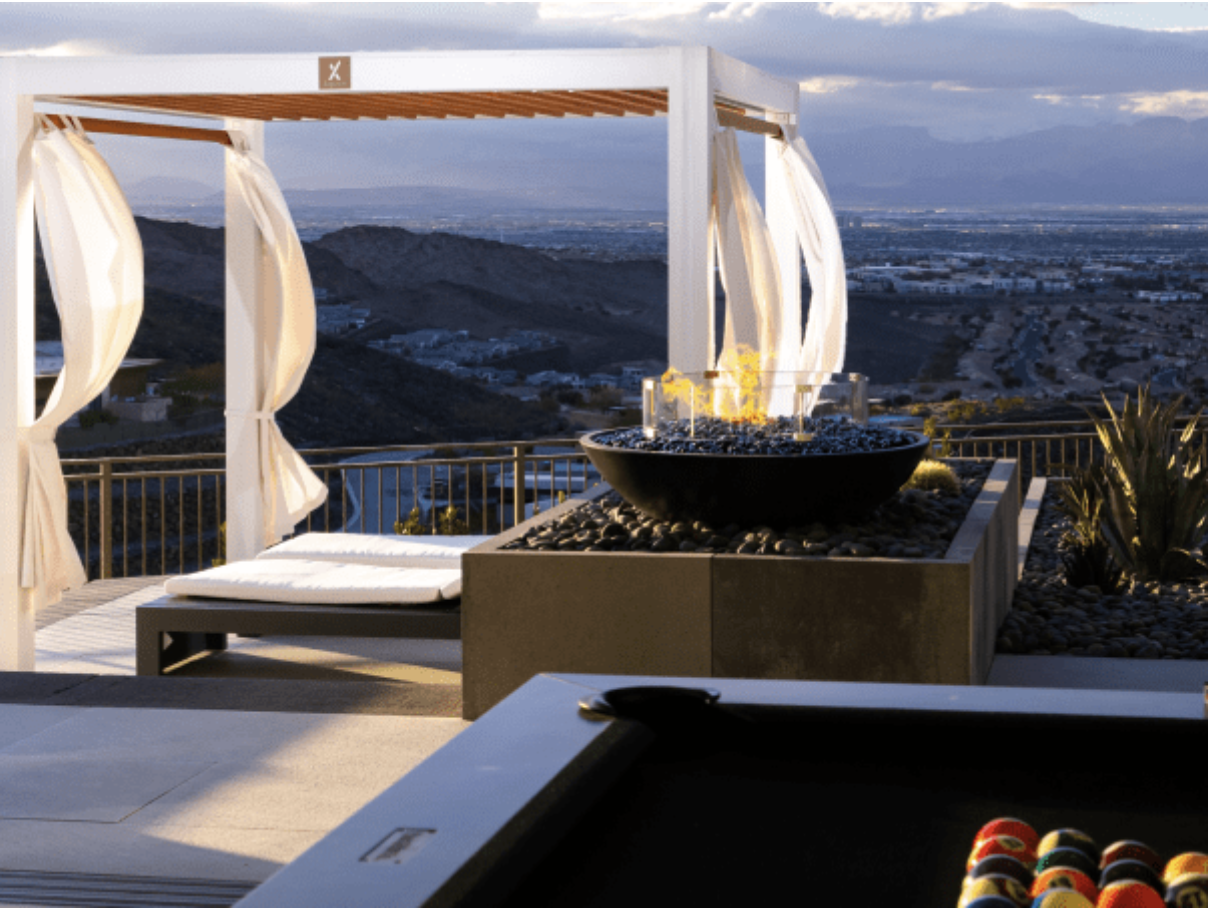Outdoor patio with pergola, fire pit, daybed, and billiards table overlooking a cityscape and mountains.