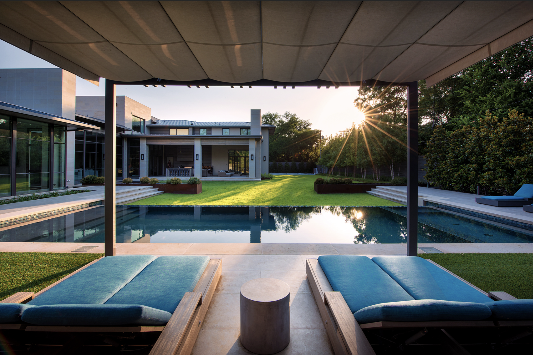 Outdoor swimming pool with lounge chairs under a canopy, modern house in the background, and sun setting over a landscaped garden.