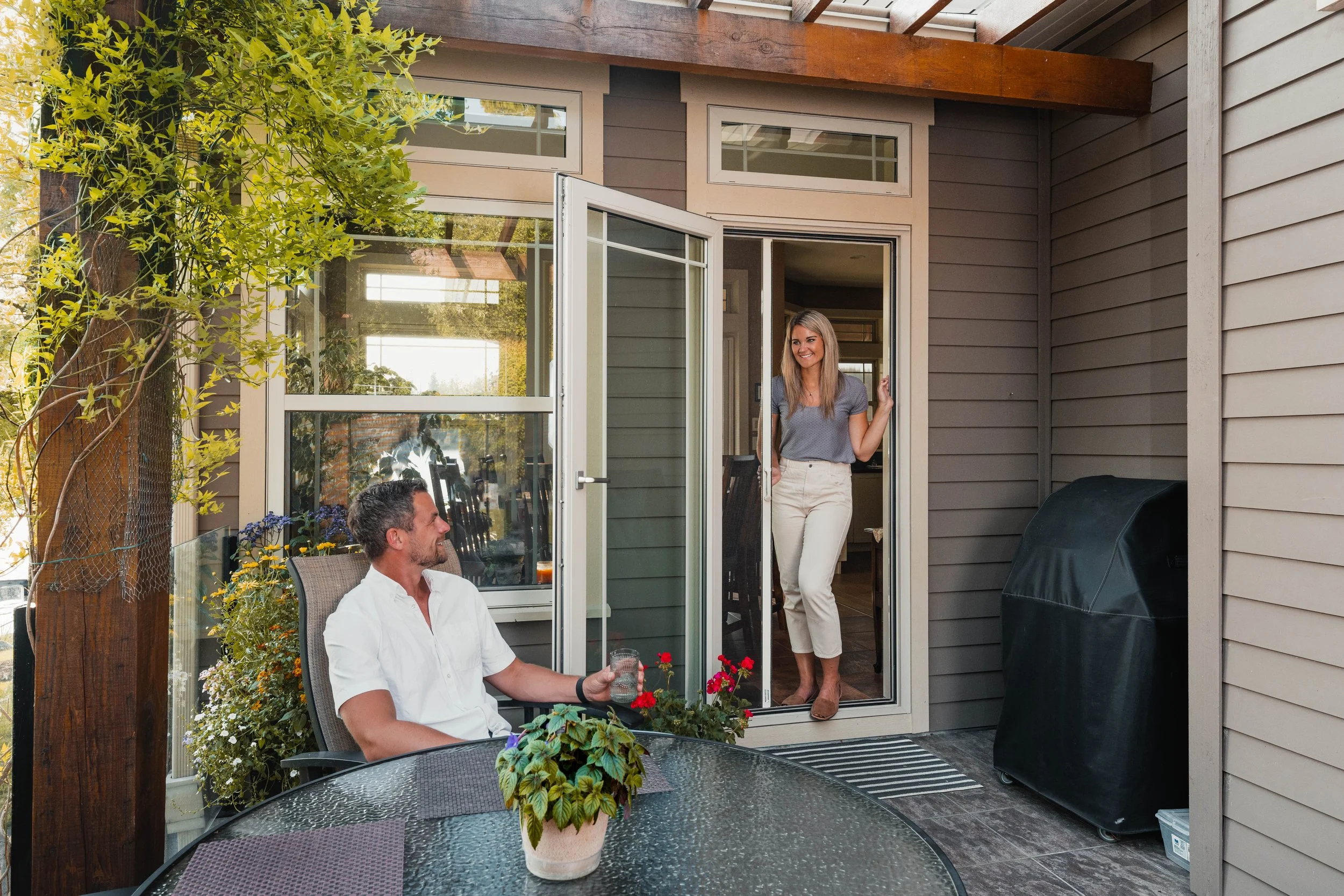 A woman standing in a doorway smiling at a man seated at a patio table with plants and a barbecue grill nearby.