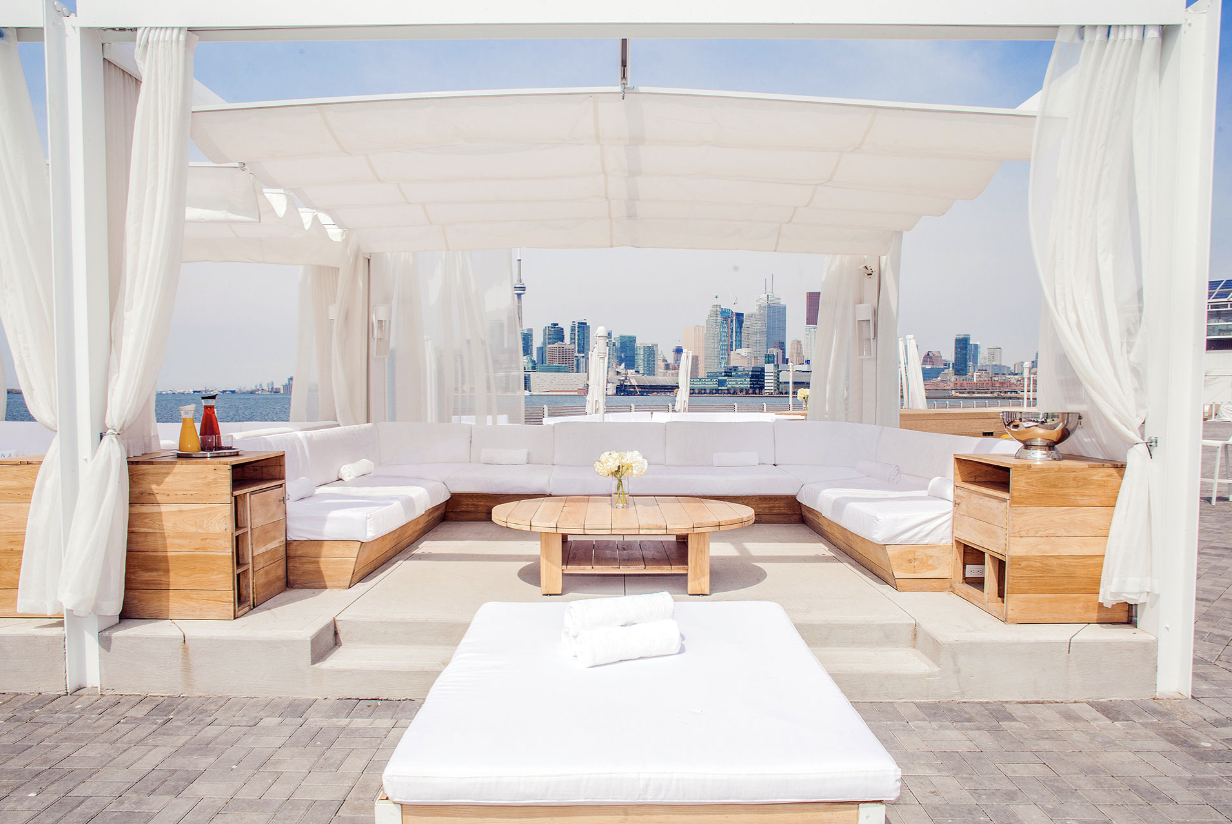 Outdoor cabana with white lounge seating, wooden furniture, and a view of a skyscraper skyline. A small table in the center holds a vase with white flowers, surrounded by open curtains.