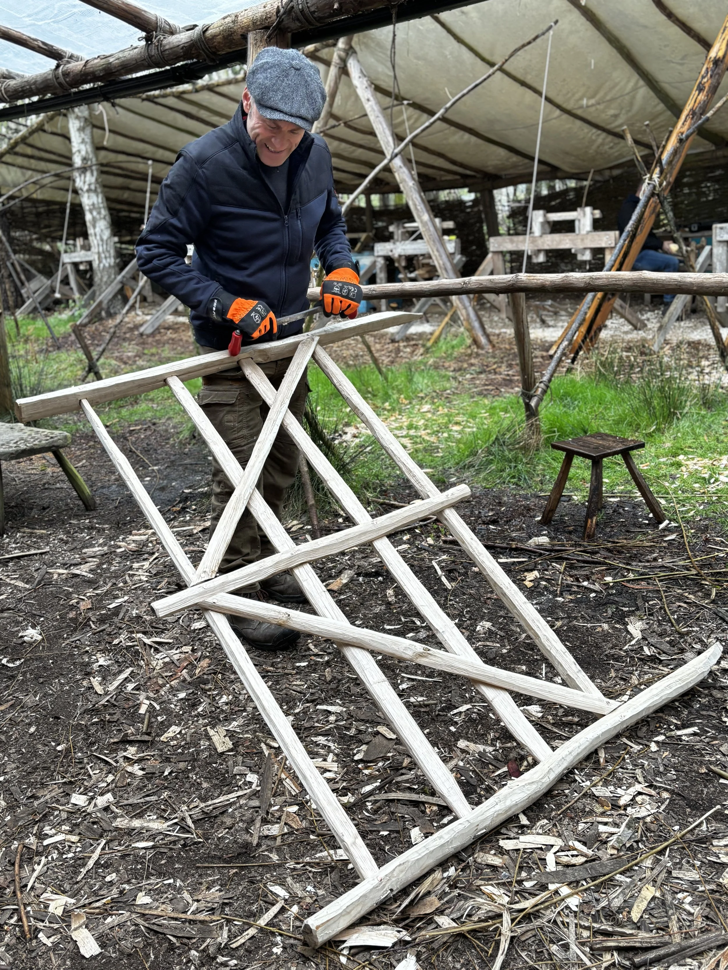 A man wearing a flat cap, black jacket, and orange gloves is sawing a piece of wood outdoors under a rustic shelter made of logs and fabric. The ground is covered with wood shavings and debris, and there are two small wooden stools near him.