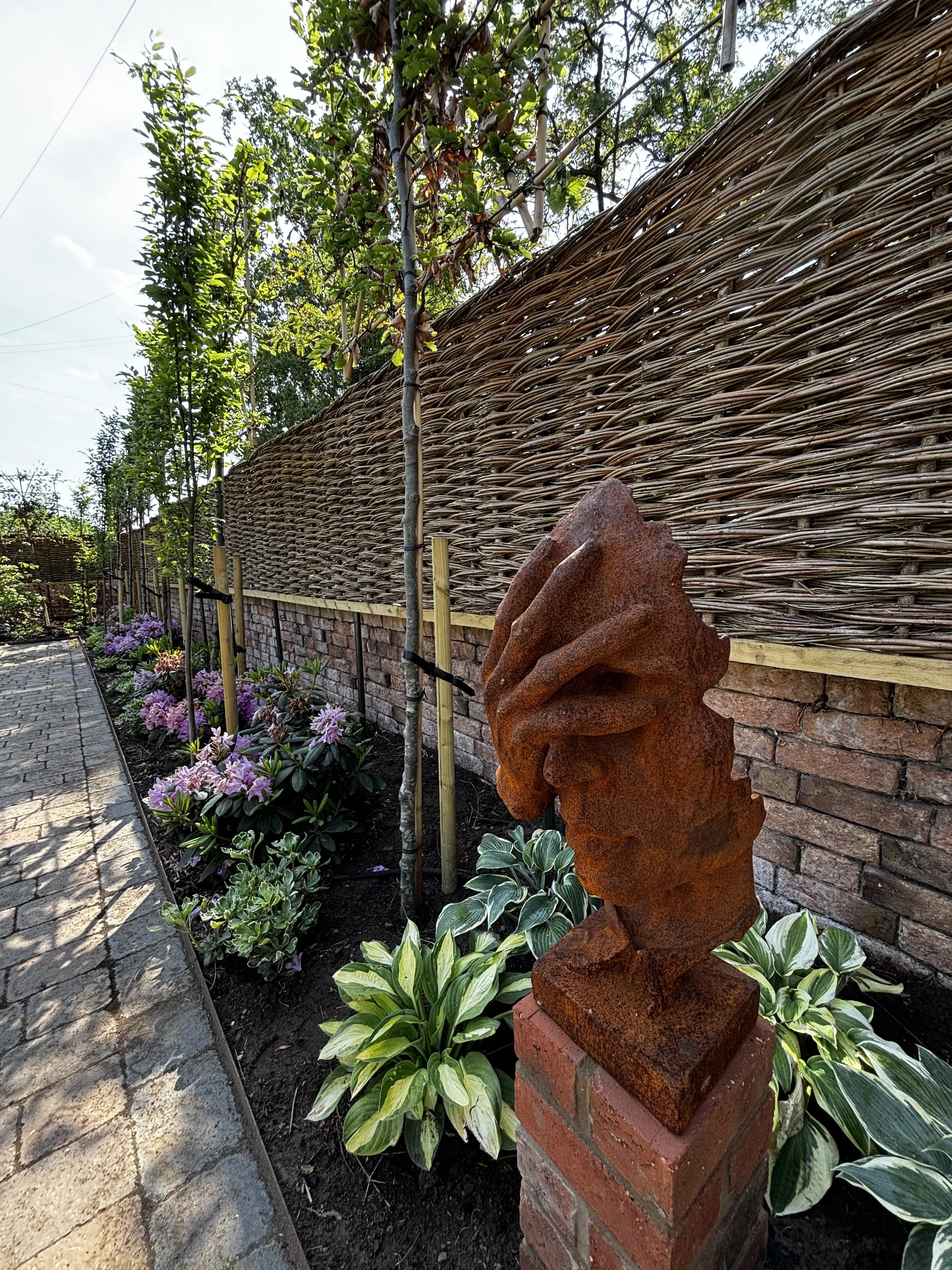 A garden scene with a brick and woven wood fence, a rusty metal sculpture, blooming pink and purple flowers, and various green plants along a cobblestone path.
