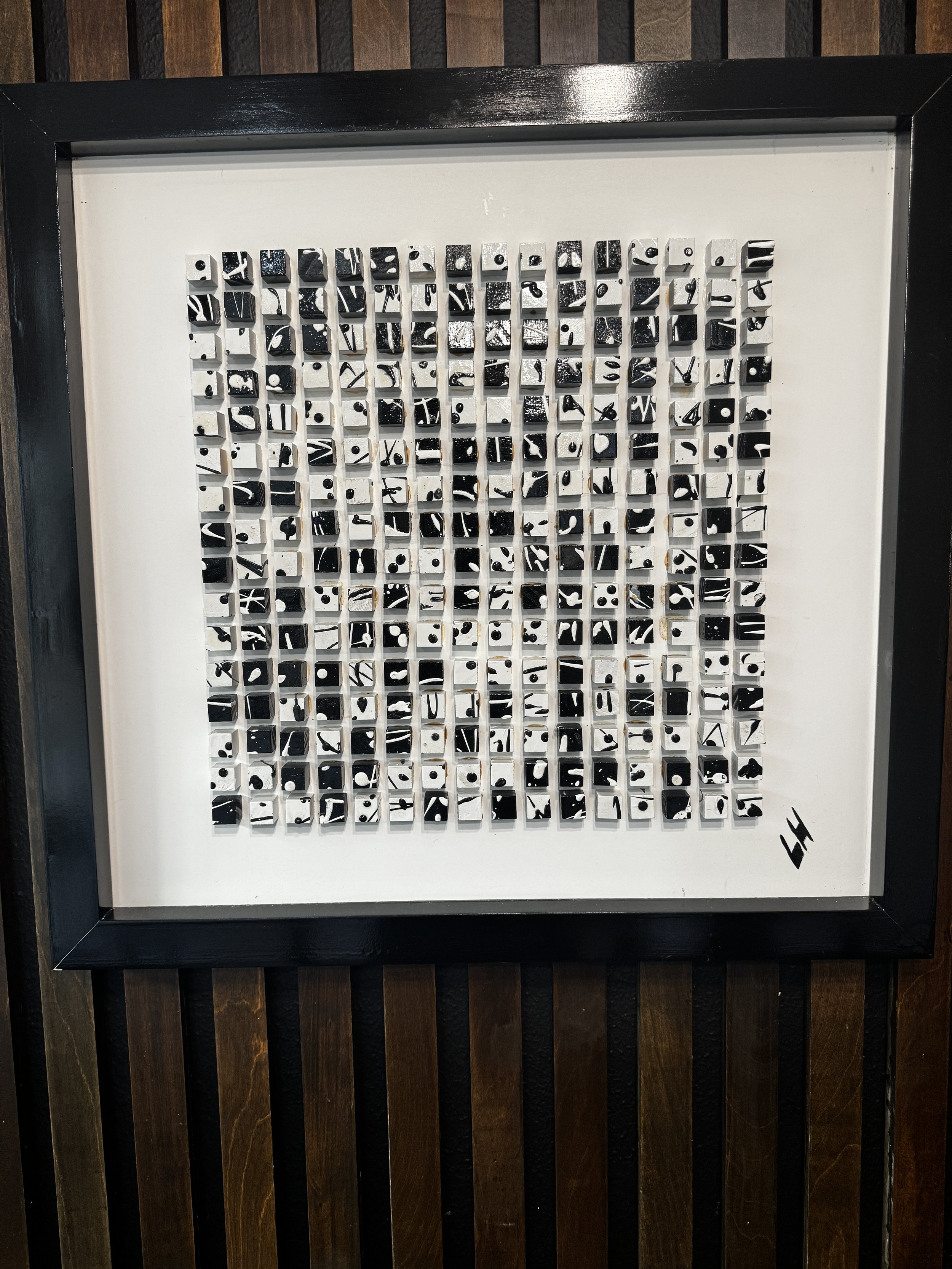 A framed mosaic artwork composed of small black and white tiles arranged in a grid pattern. The mosaic is set against a white background, with a black frame surrounding it. The artwork has an abstract design with various black and white shapes and li