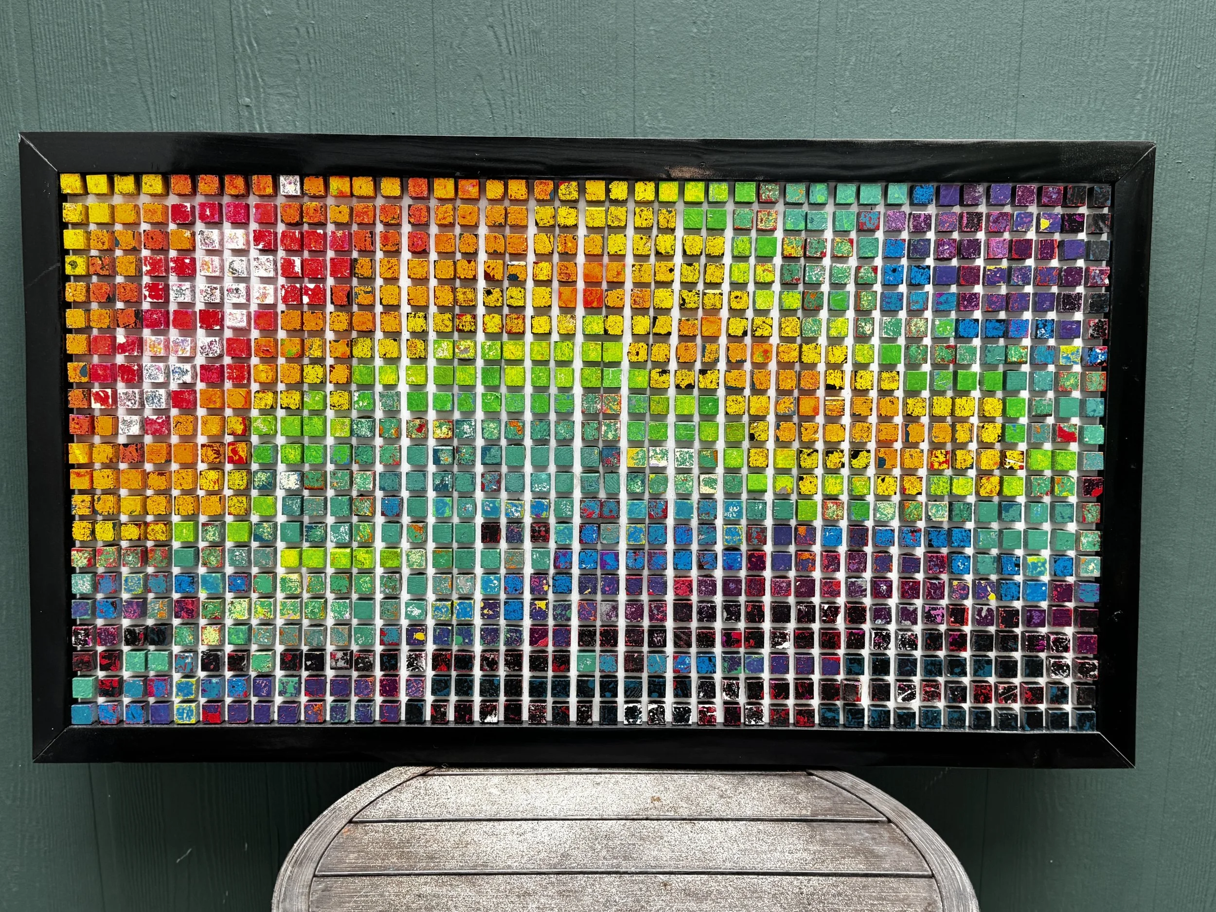 Colorful mosaic artwork with small square tiles arranged in rainbow gradient, framed in black, placed on a green wall above a wooden bench.