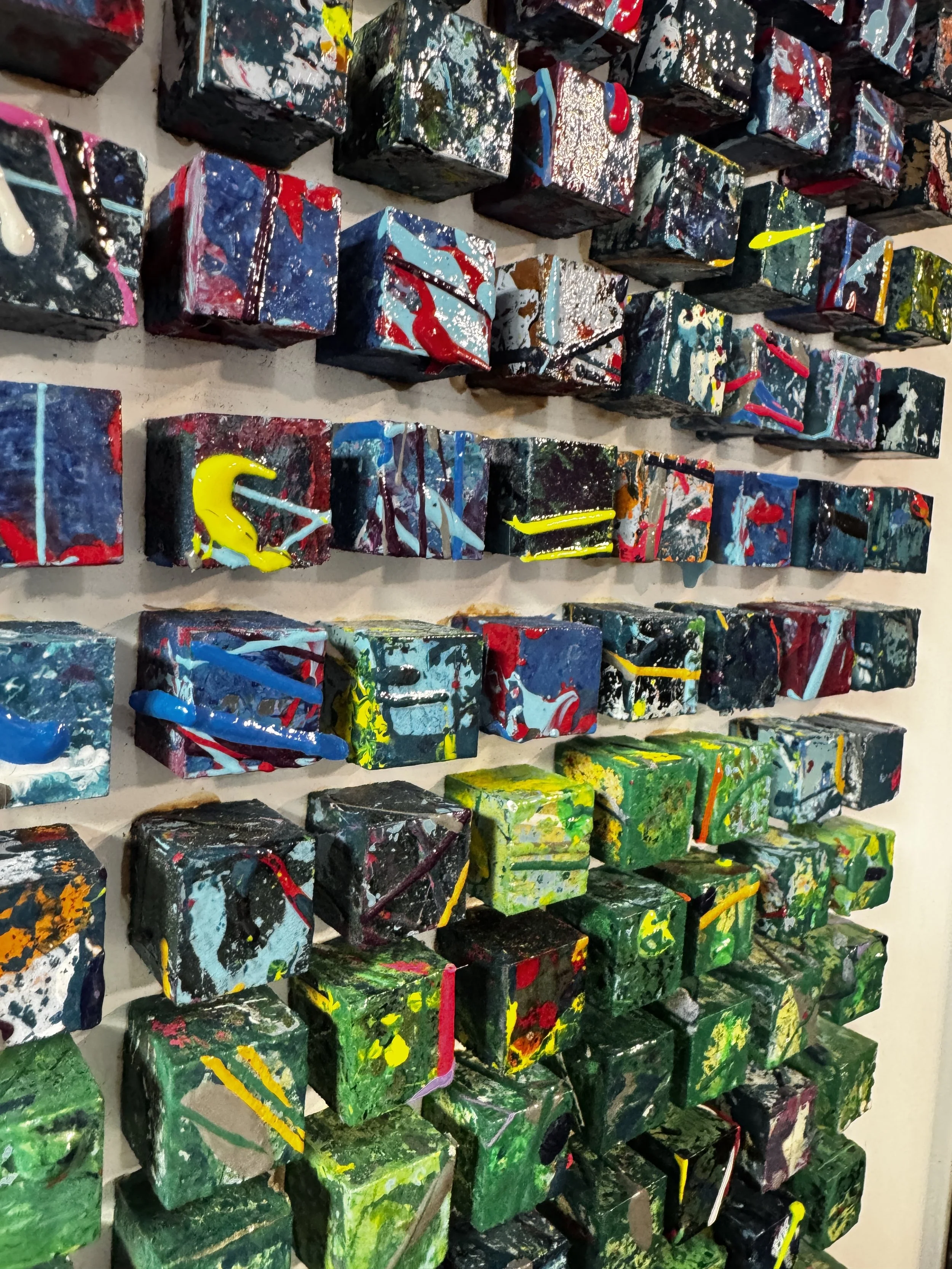 A wall display of small, colorful, abstract painted cubes arranged in a grid pattern.
