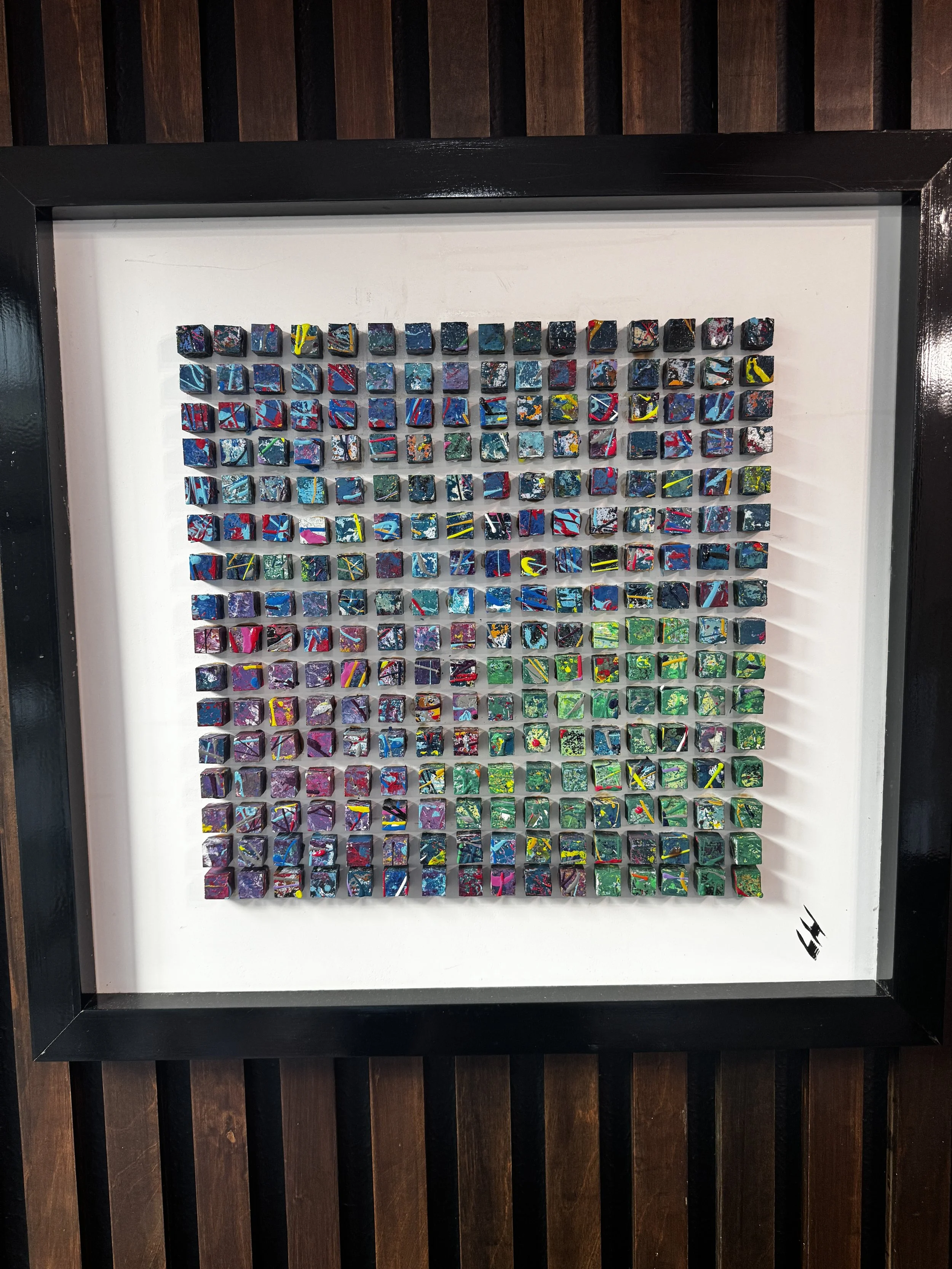 Colorful abstract mosaic art with small square tiles arranged in a grid, displayed in a black frame on a wooden wall.