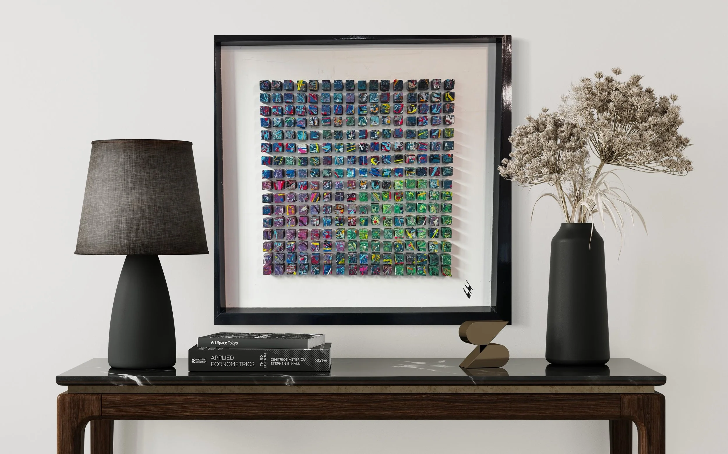 Decorative wall artwork consisting of colorful, abstract square pieces framed in black, displayed on a white wall above a black marble-topped wooden console table. The table holds a black table lamp with a gray shade, several books including "Applied