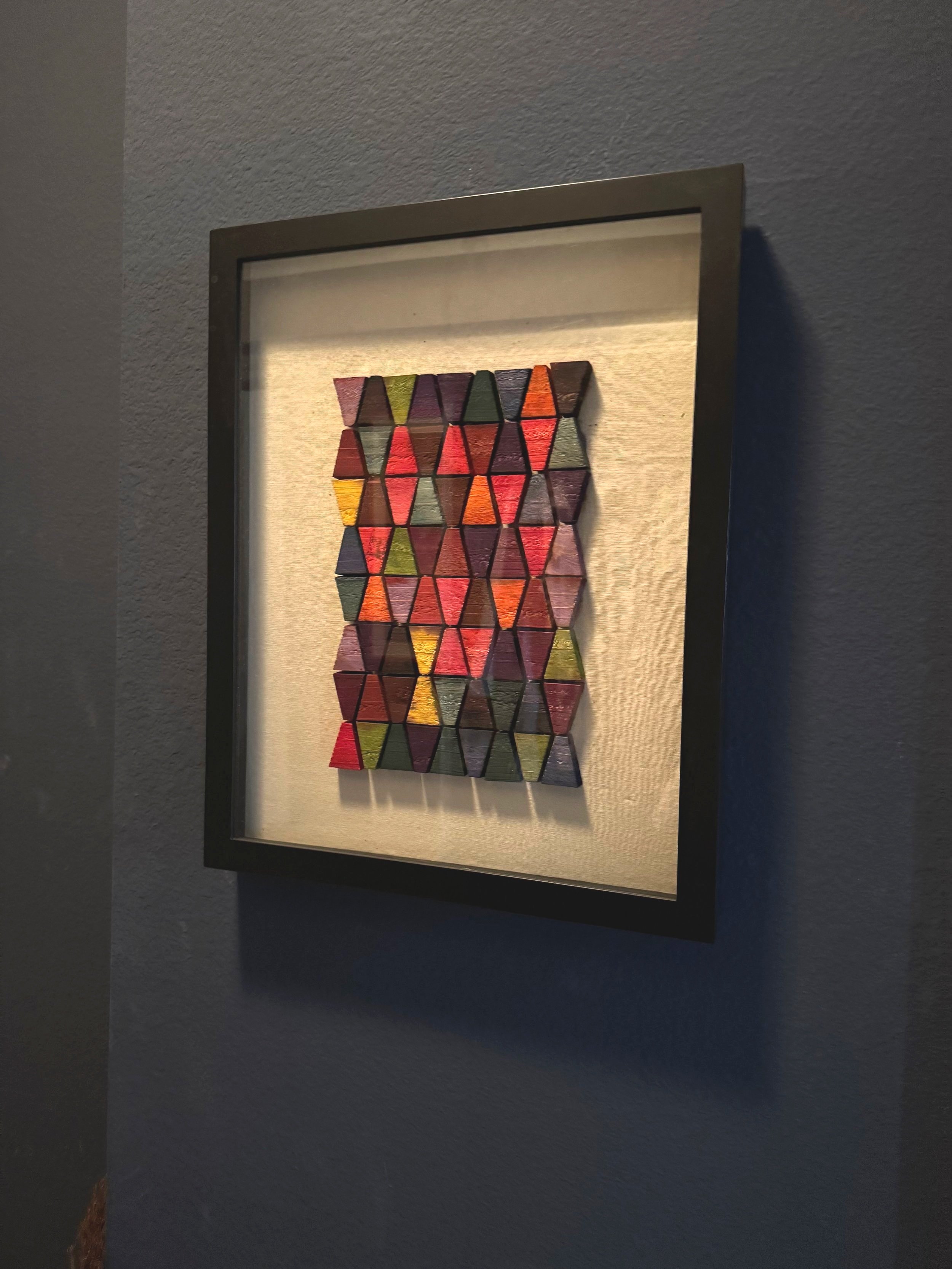 A framed artwork featuring a pattern of small, colorful, triangular wooden blocks arranged in a grid on a beige background, hanging on a gray wall.