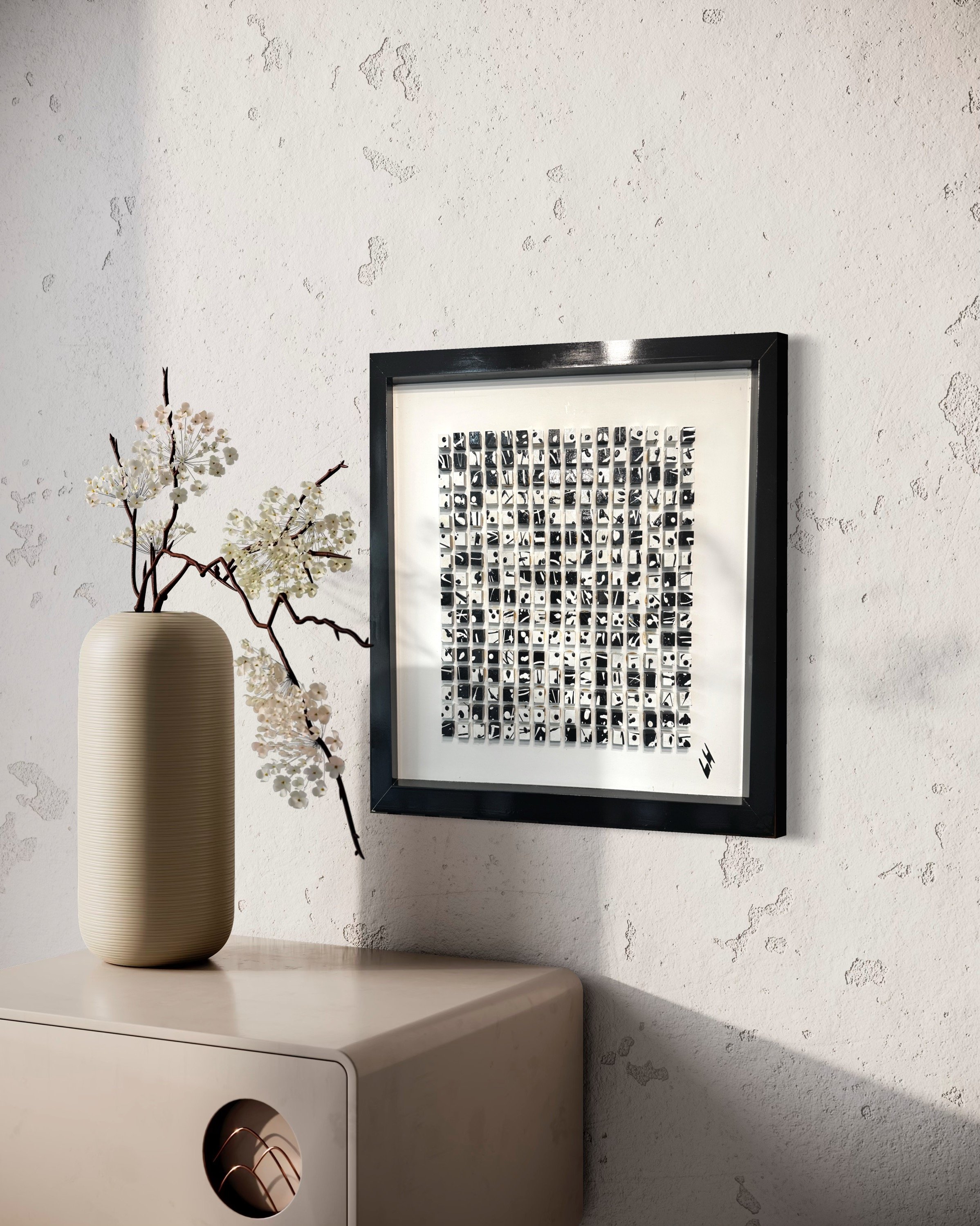 A beige vase with white flowers and dark branches sits on a beige cabinet, next to framed black and white artwork on a textured white wall.