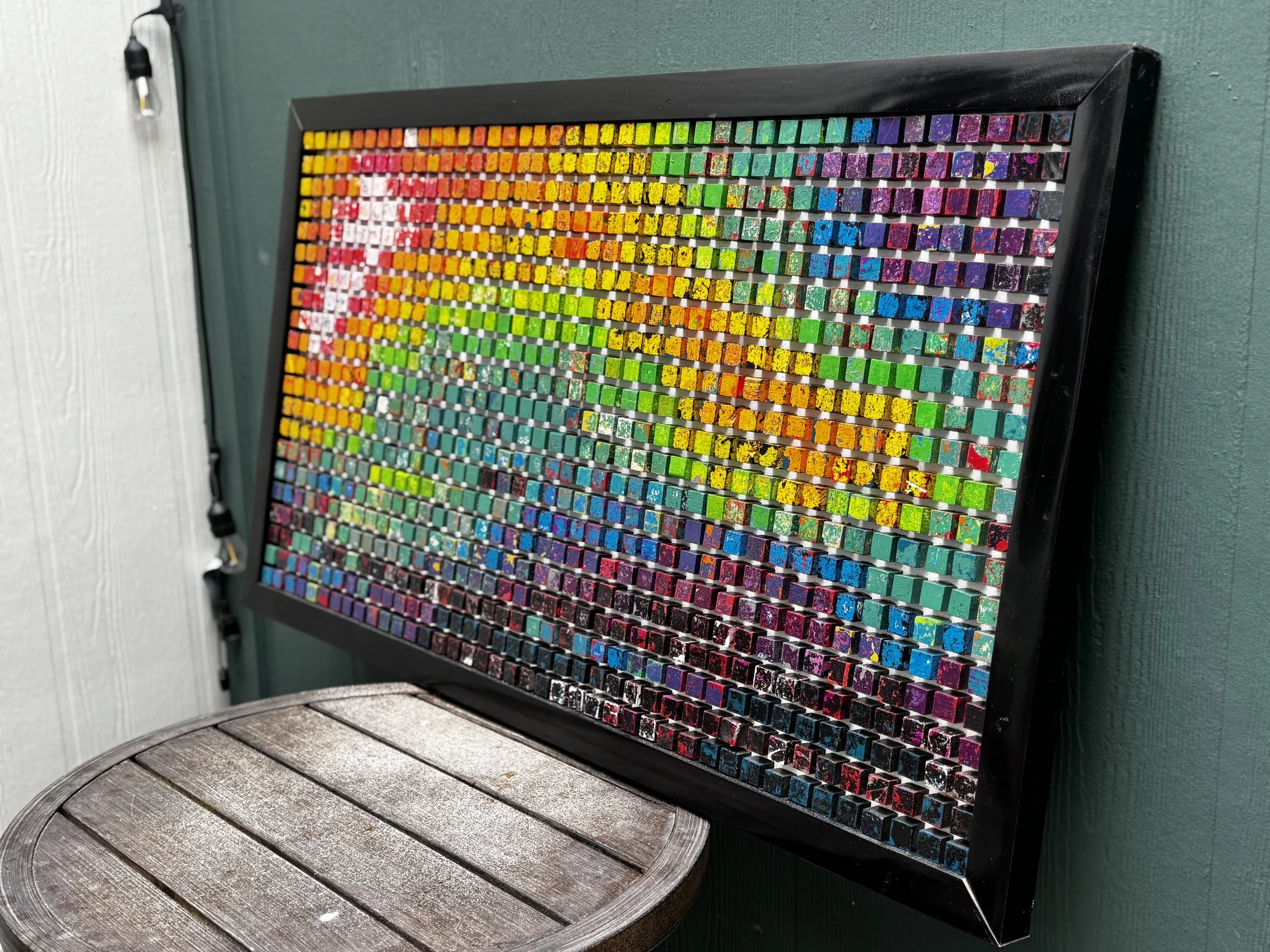 Colorful mosaic art piece with small, square, glass tiles in various vibrant colors, framed in black, leaning against a teal wall.
