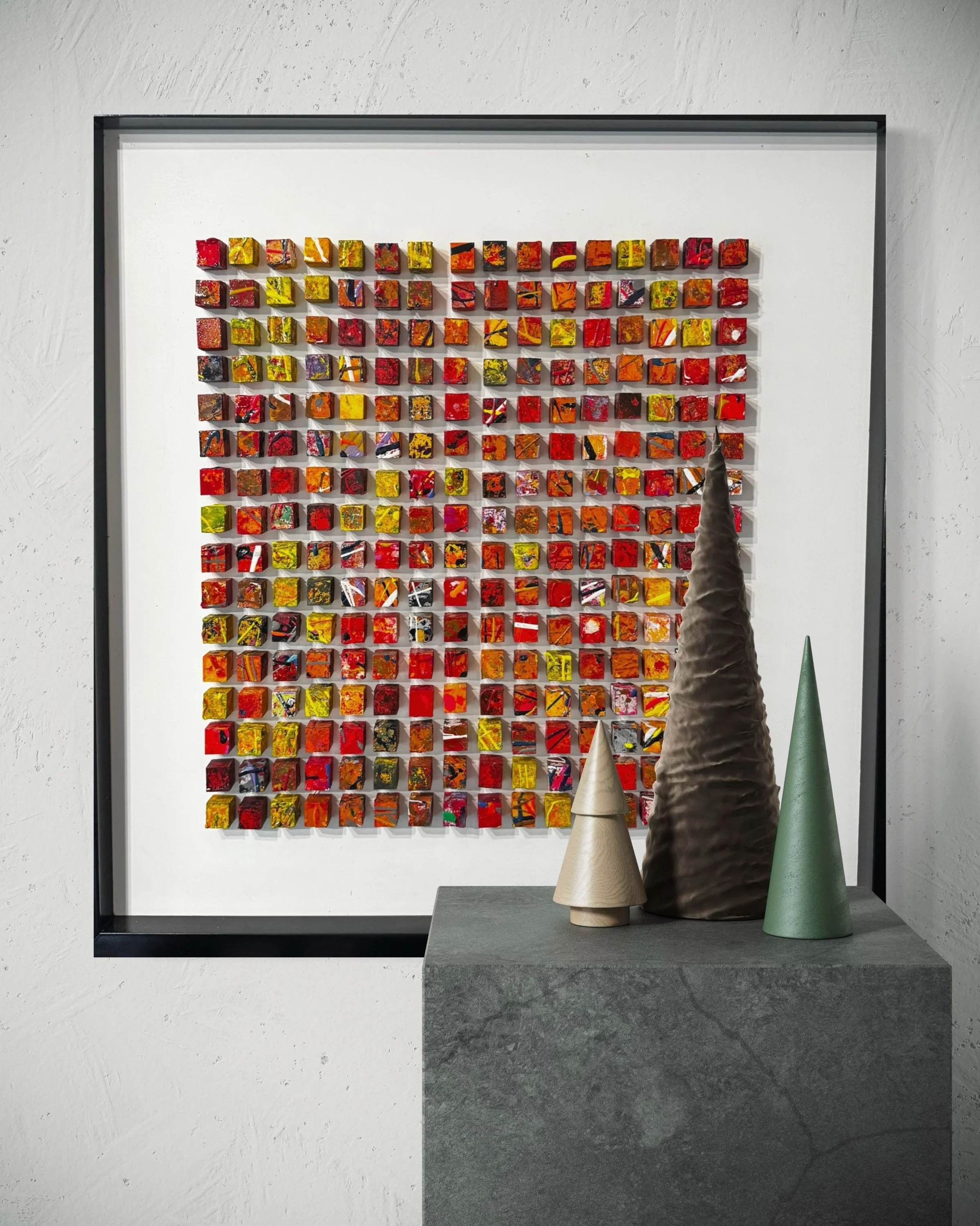 A framed artwork composed of a grid of small, colorful, abstract tiles and three decorative cone-shaped objects on a gray pedestal in front of it.