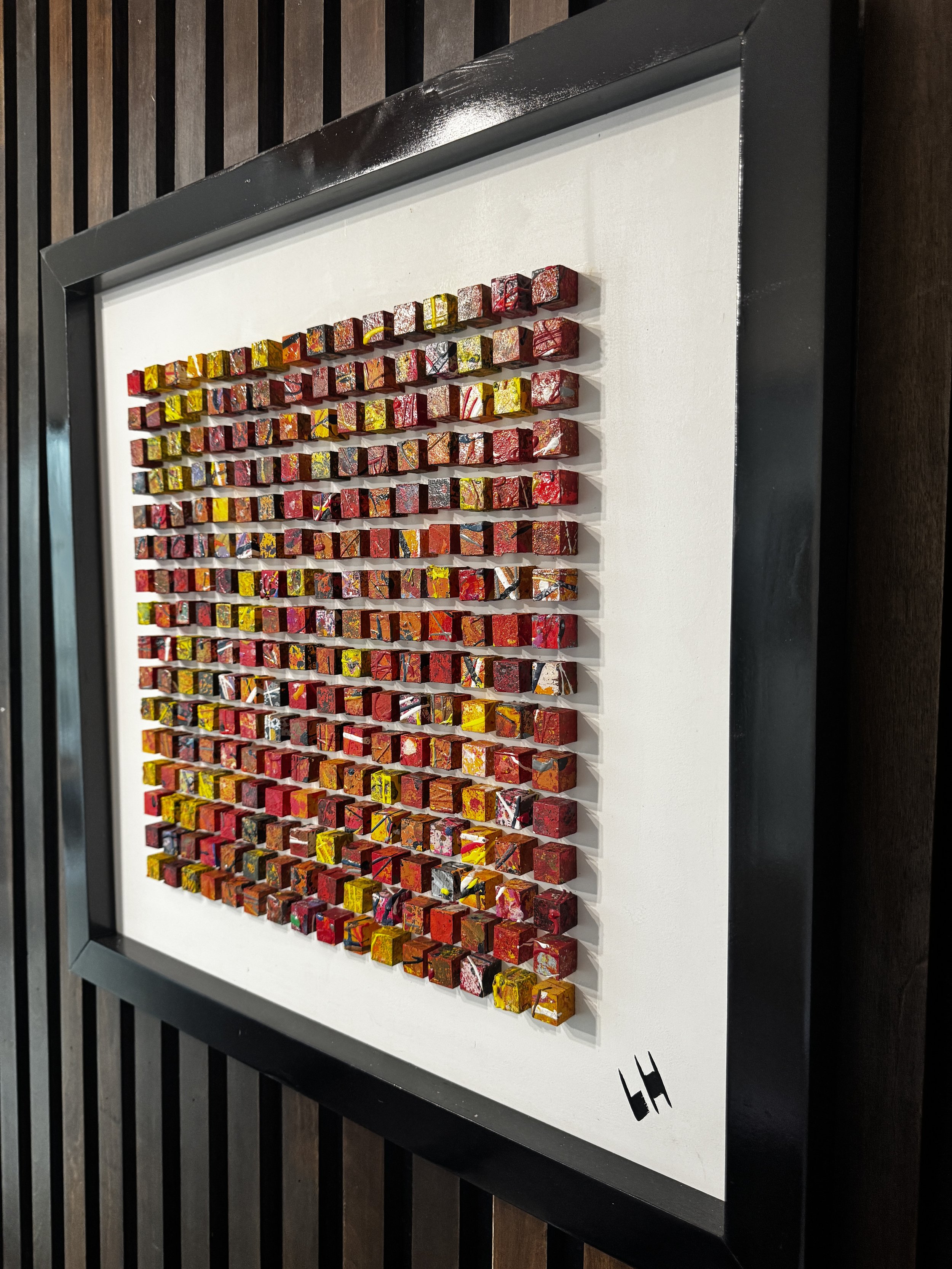 A framed mosaic artwork with numerous small, colorful, cube-shaped tiles arranged in a grid pattern on a white background, mounted on a dark wooden wall.