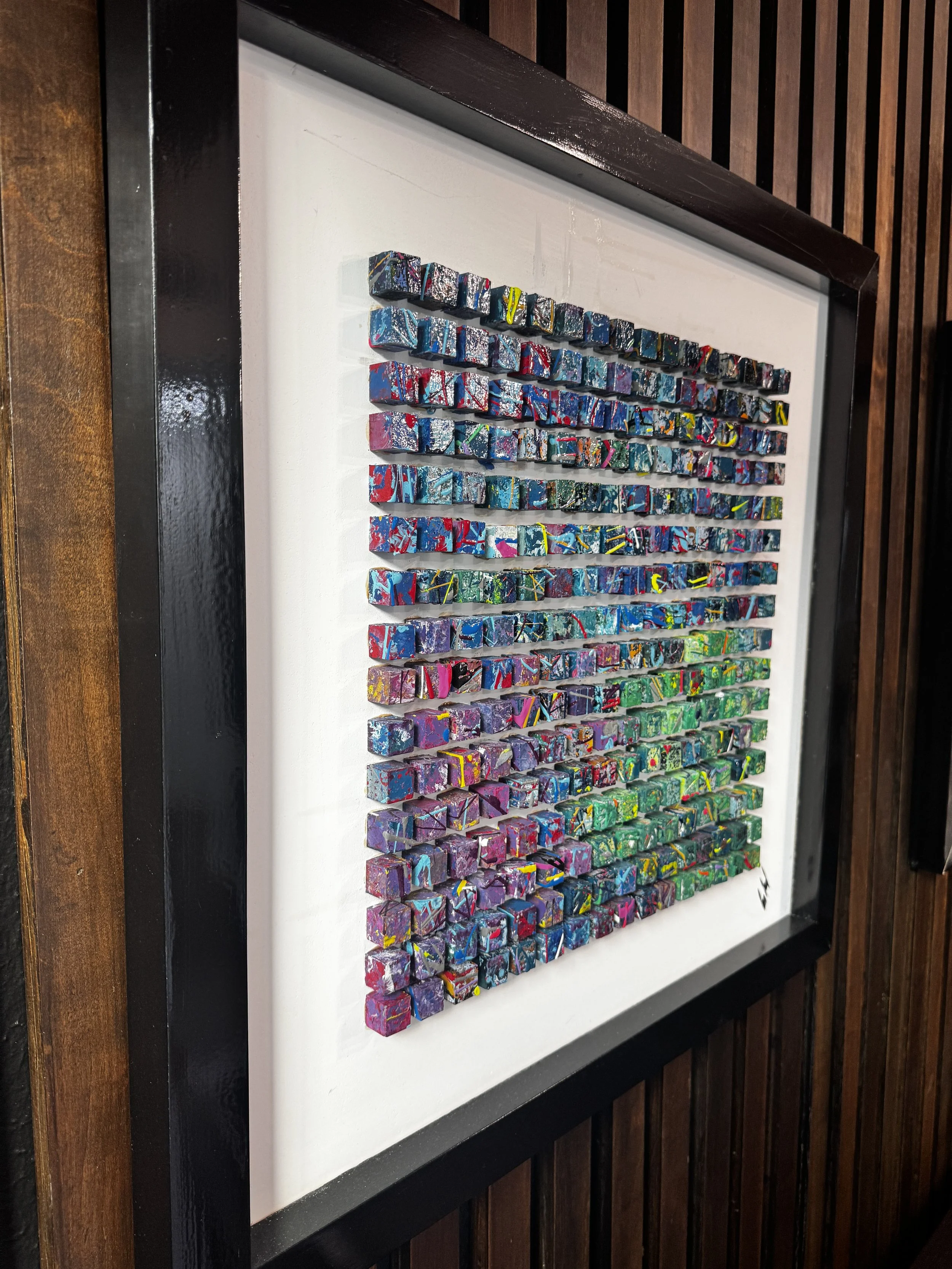 Colorful abstract mosaic artwork with small square tiles arranged in a grid, mounted in a black frame on a wall with wooden paneling.