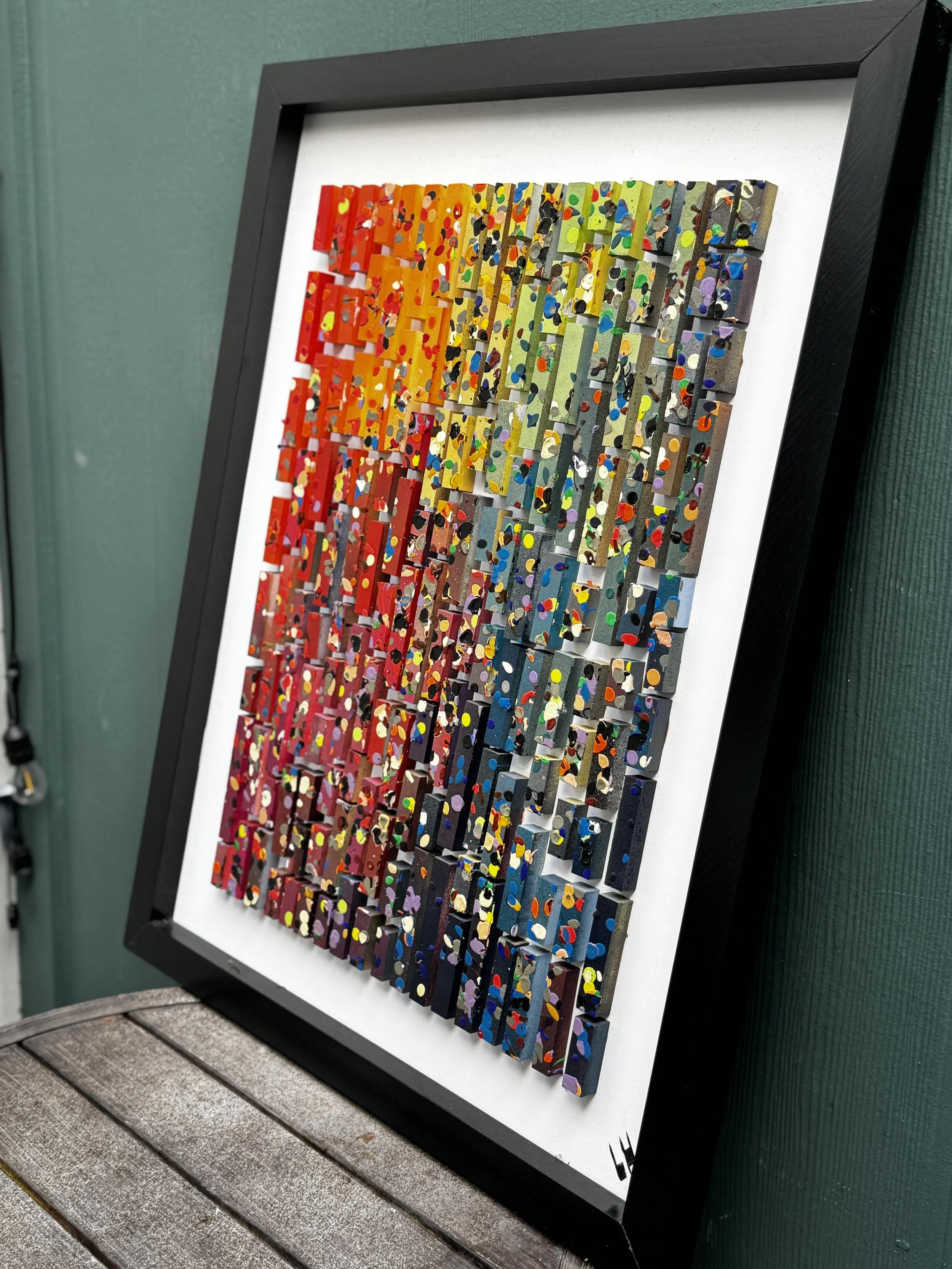 Colorful abstract artwork with multicolored dots and shapes on rectangular pieces, arranged in a grid within a black frame, leaning against a green wall.