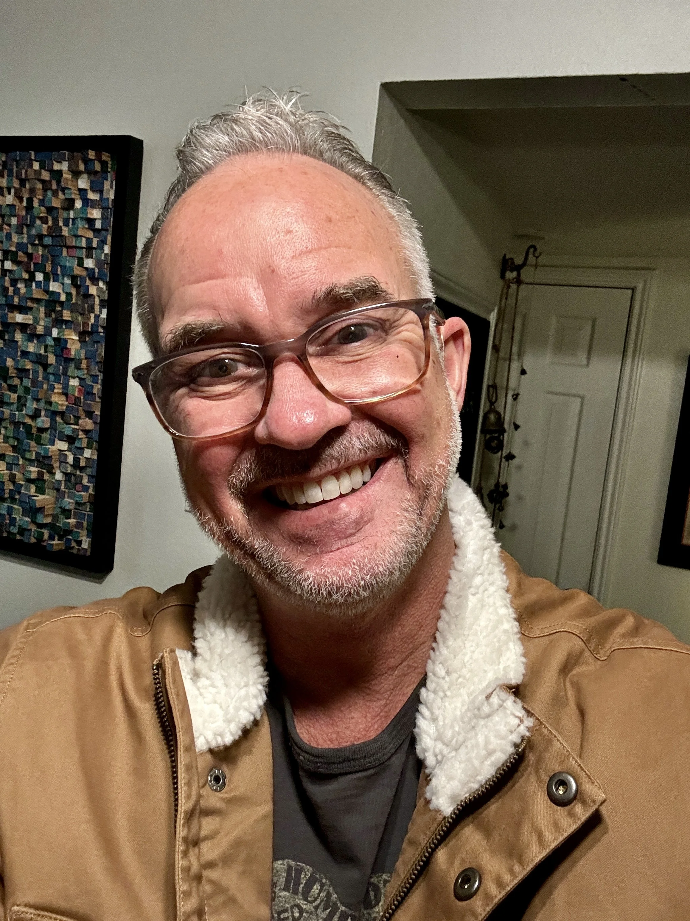 A smiling middle-aged man with short gray hair, glasses, and a beard, wearing a brown jacket with a white fleece collar. He is indoors with artwork on the wall behind him and a door in the background.