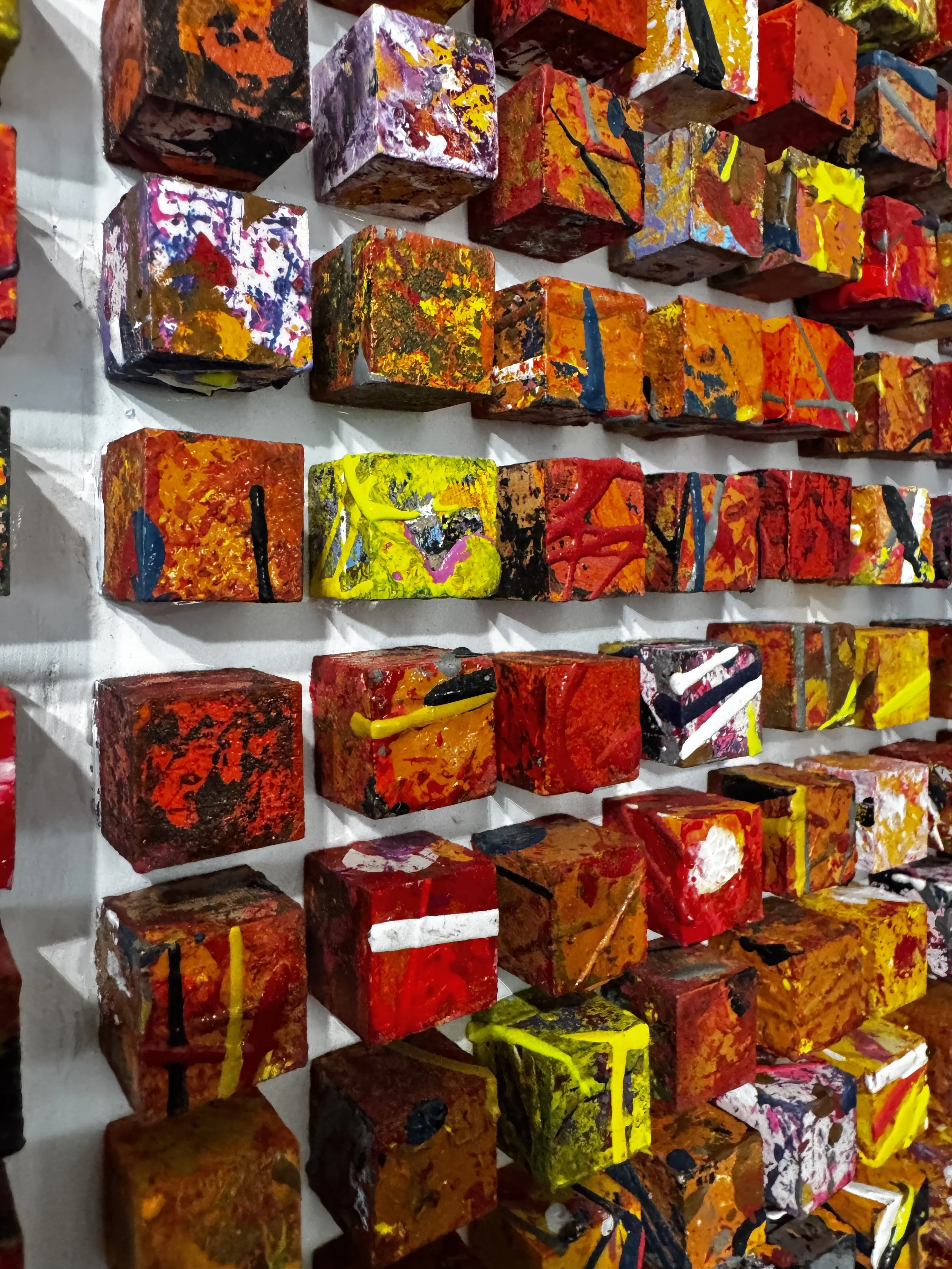 Close-up of a wall with multiple small, colorful, abstract painted blocks arranged in rows.