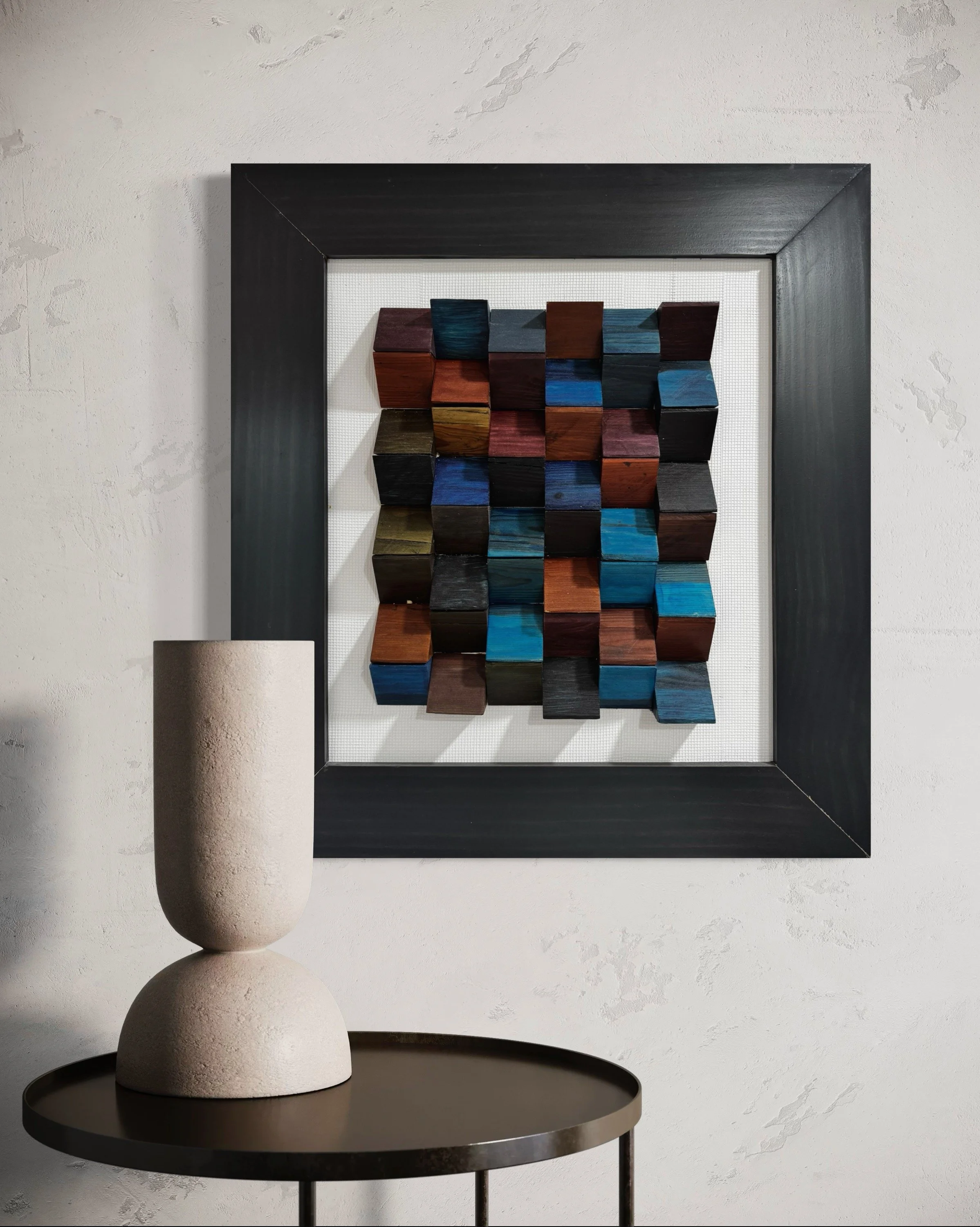 Artwork of multi-colored wooden cubes mounted on a white wall, framed with black trim, with a beige textured vase on a round black table in front.
