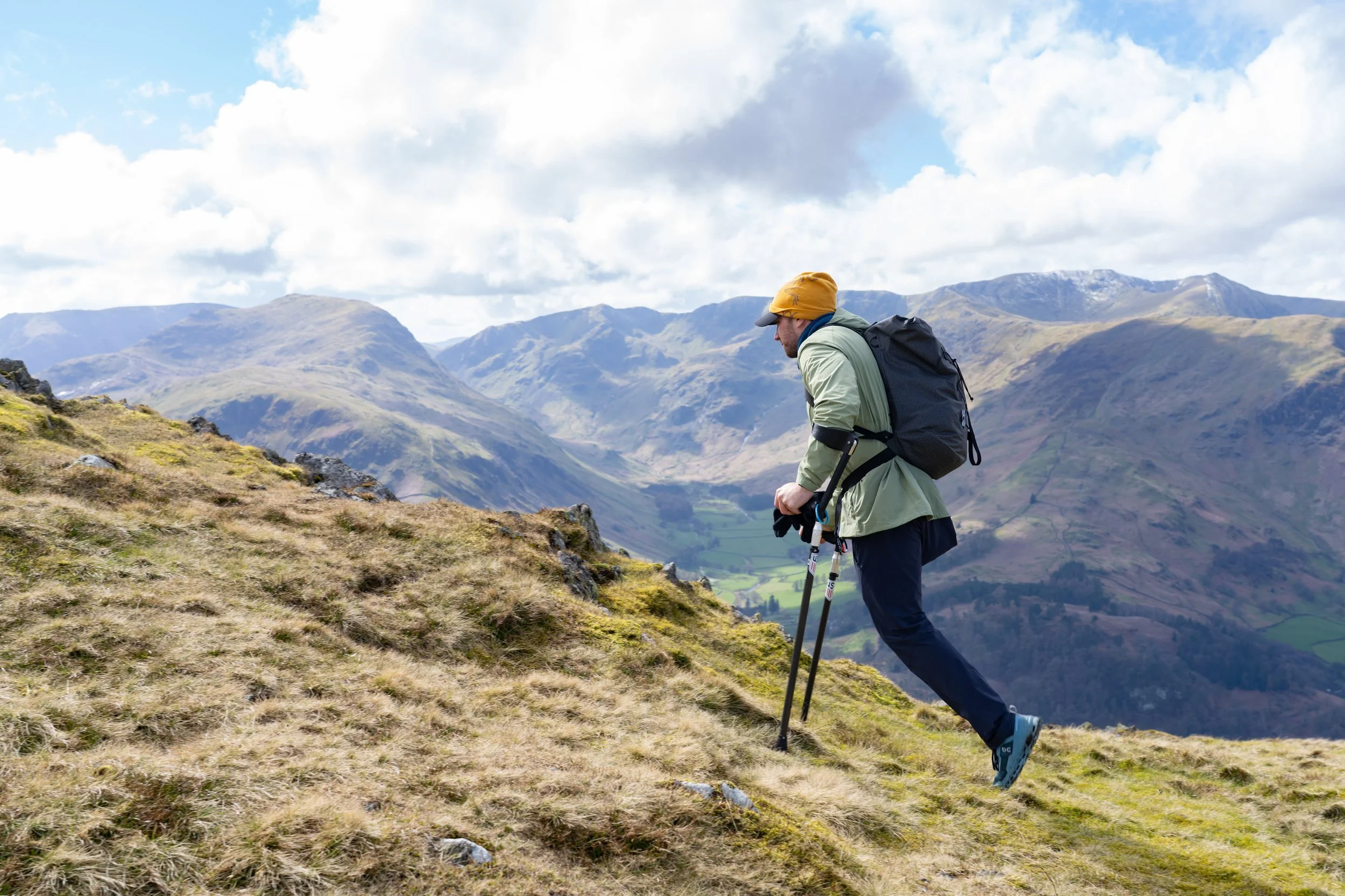 Hiker with walking poles on grassy mountain trail, wearing a backpack and yellow beanie, against a backdrop of mountains and cloudy sky.