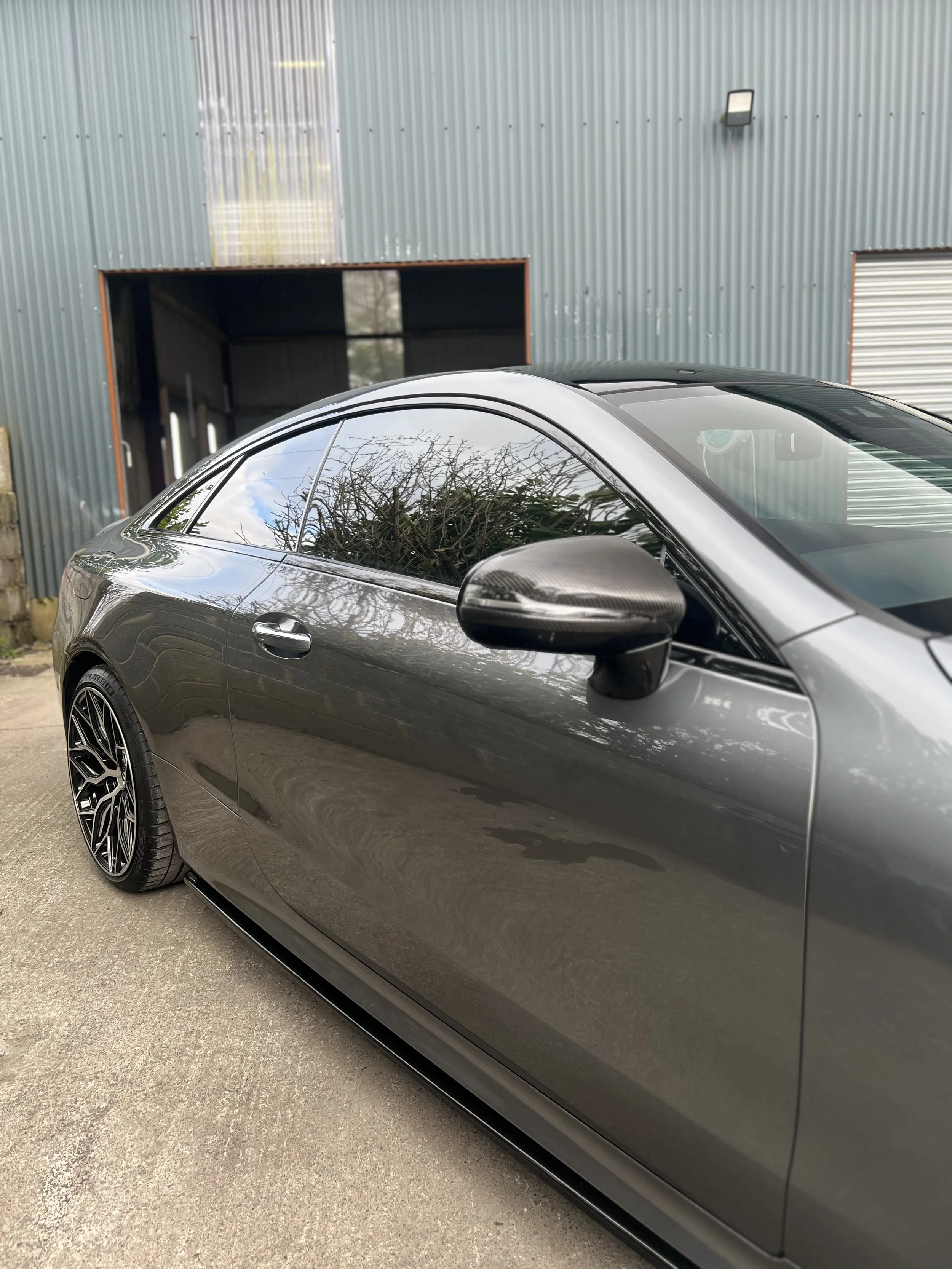 Side view of a gray sports car parked outside a metal building with a garage door and a roll-up door.