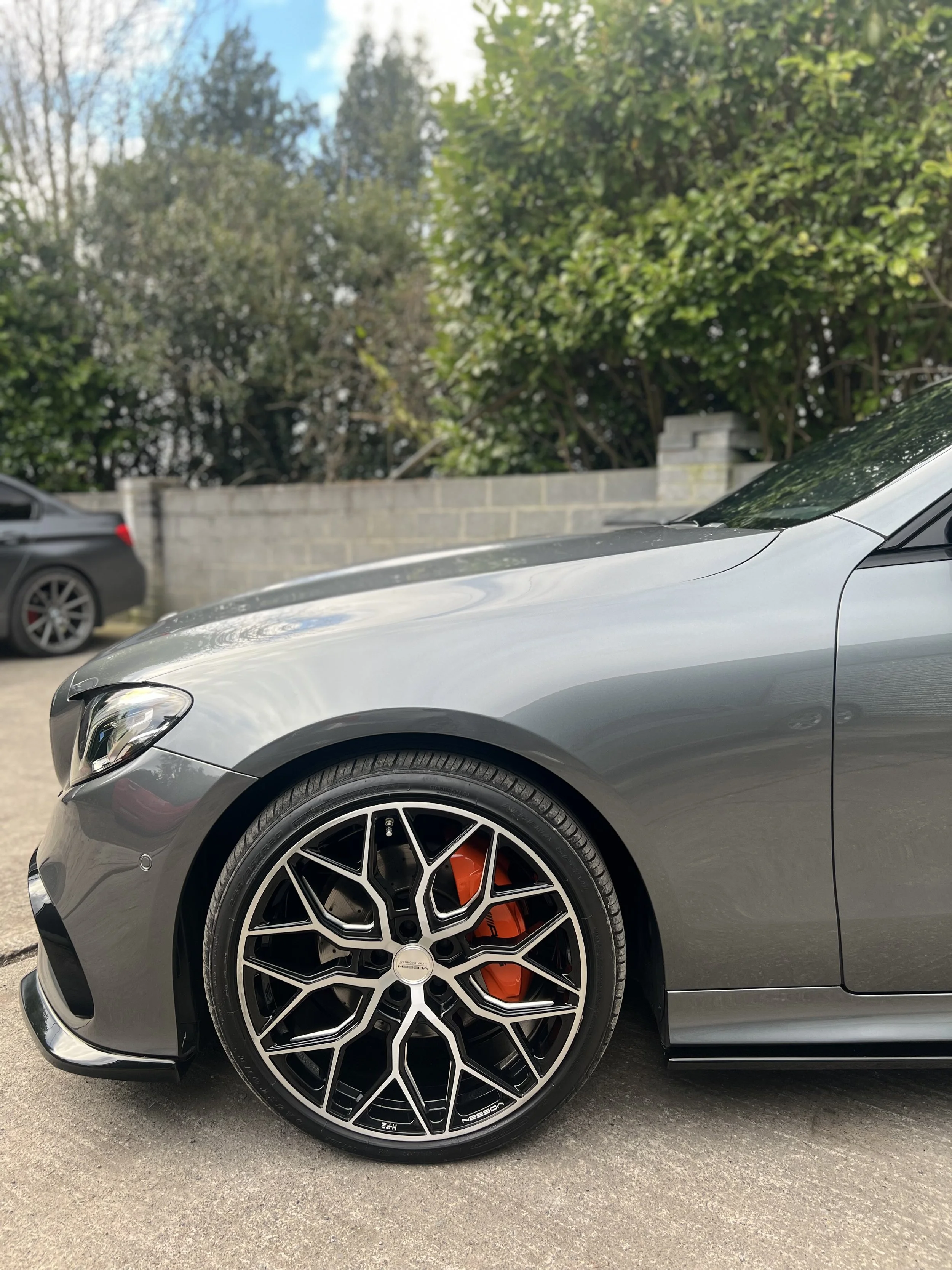 Close-up of the front part of a silver sports car with black and silver alloy wheel and orange brake caliper, parked on a paved surface with trees and a gray wall in the background.