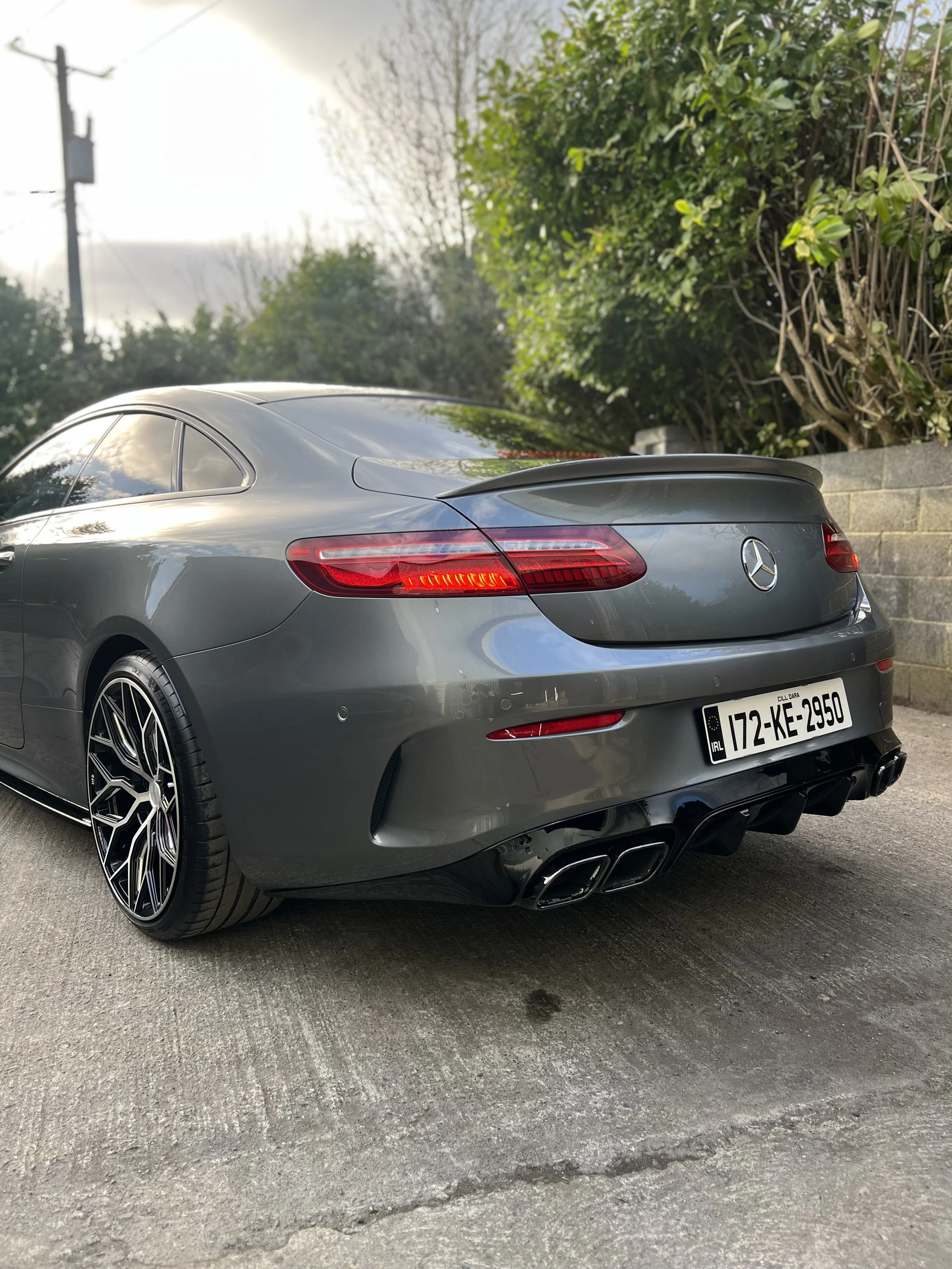 A gray Mercedes-Benz coupe parked on a road with trees and bushes in the background, showing the rear view with dual exhausts and a European license plate.