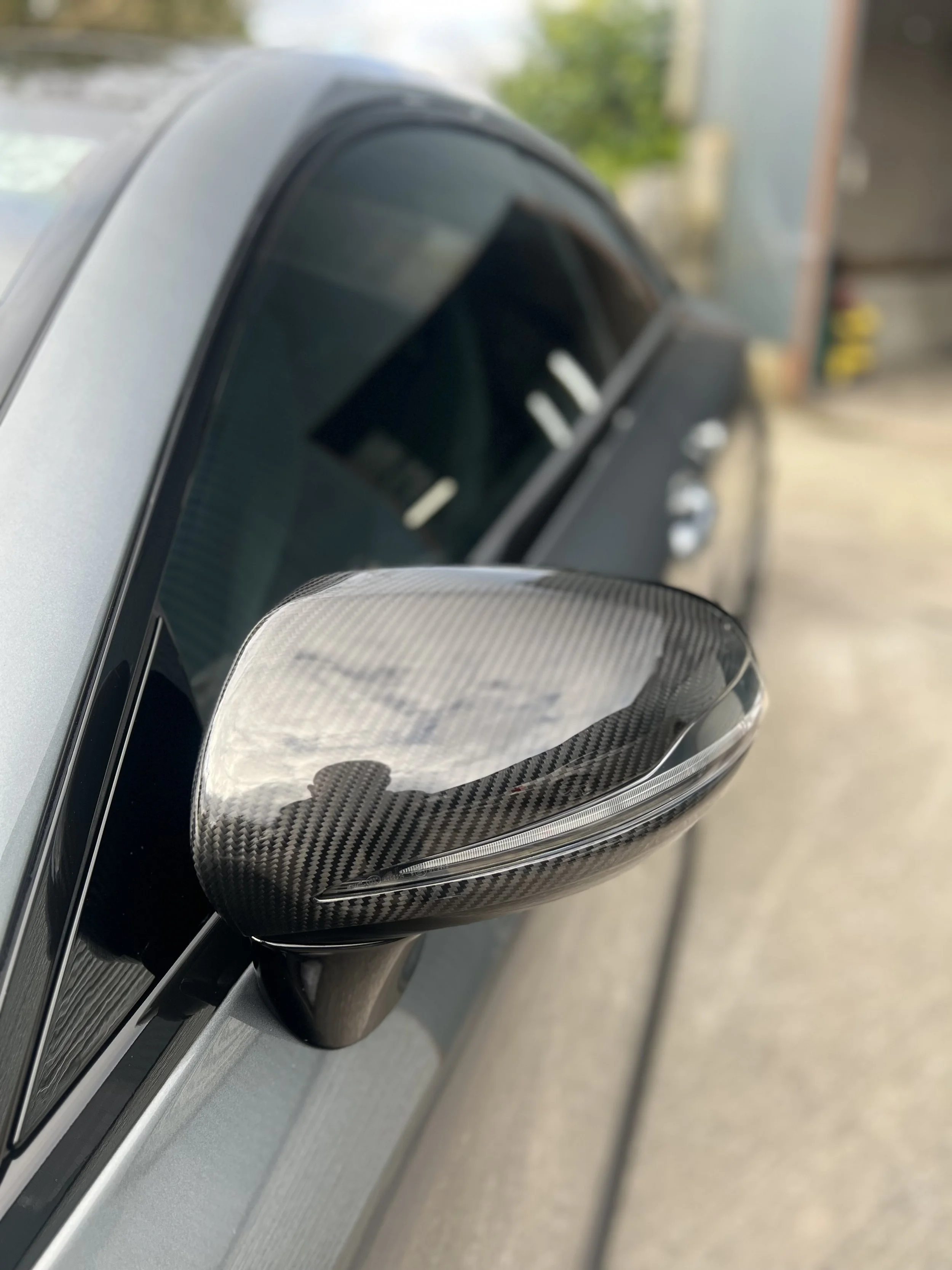Close-up of a carbon fiber side mirror on a silver car, with a blurred background showing part of a garage door and some outdoor greenery.