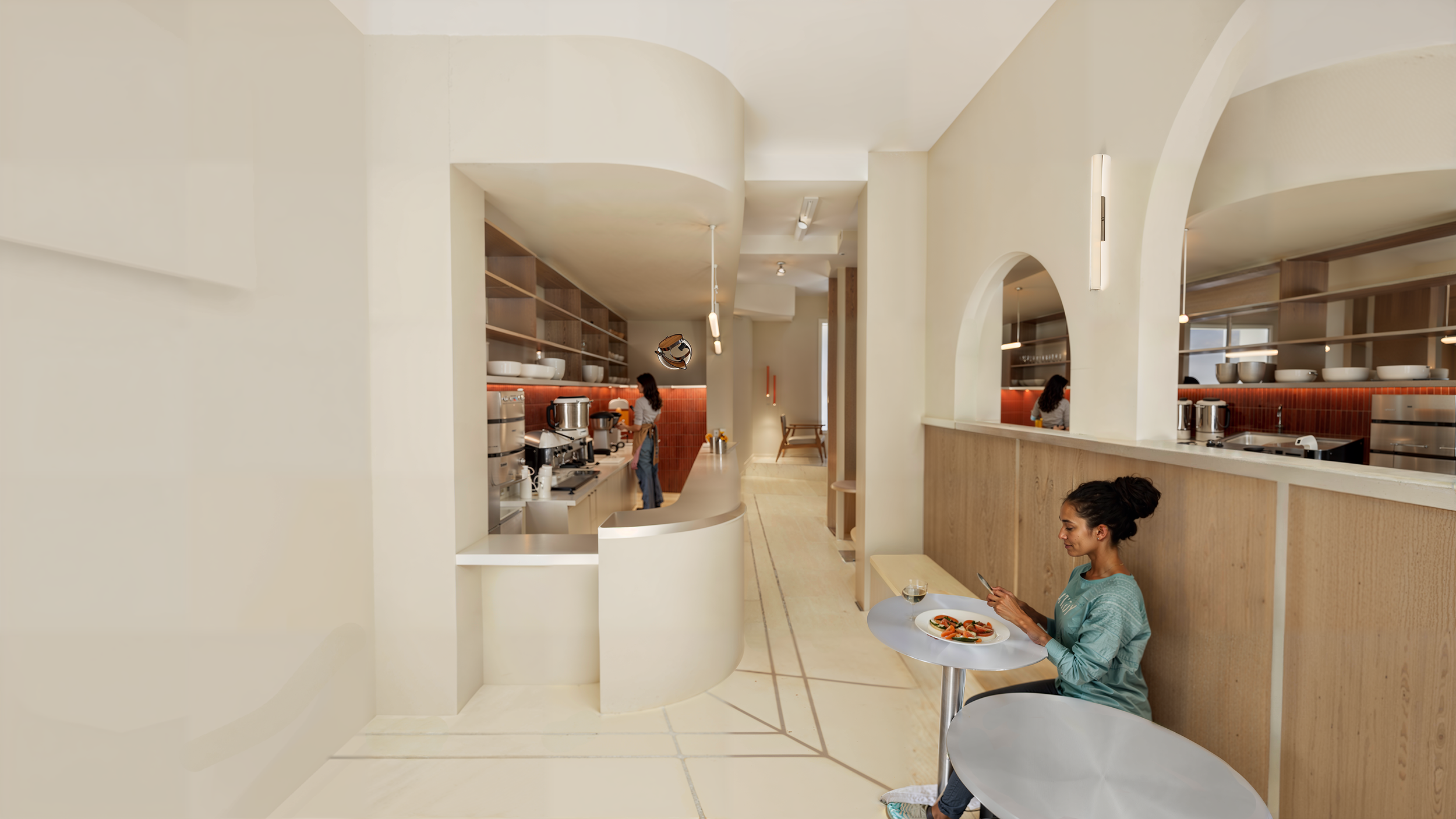 Une femme assise à une table dans un café, utilisant son téléphone, avec une assiette de nourriture devant elle. En arrière-plan, une personne travaille dans une cuisine moderne ouverte.