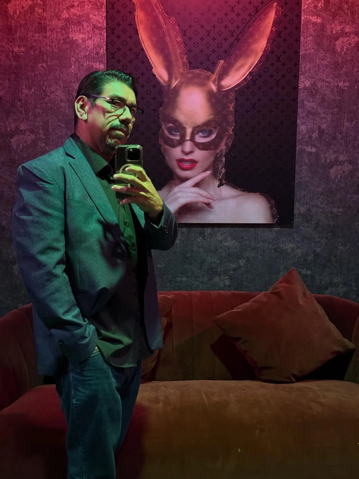 A man in a blazer taking a mirror selfie in a dimly lit room with purple and pink lighting. Behind him is a large portrait of a woman with bunny ears and a mask, featuring Louis Vuitton patterns. The room has a velvet sofa with cushions.