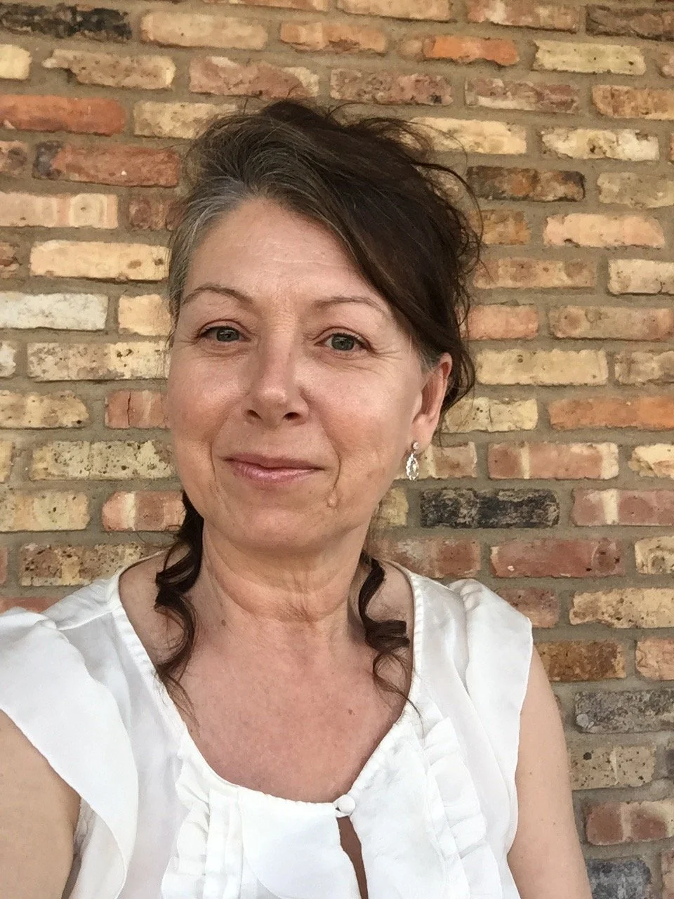 A woman with brown hair and earrings, wearing a white top, taking a selfie in front of a brick wall.