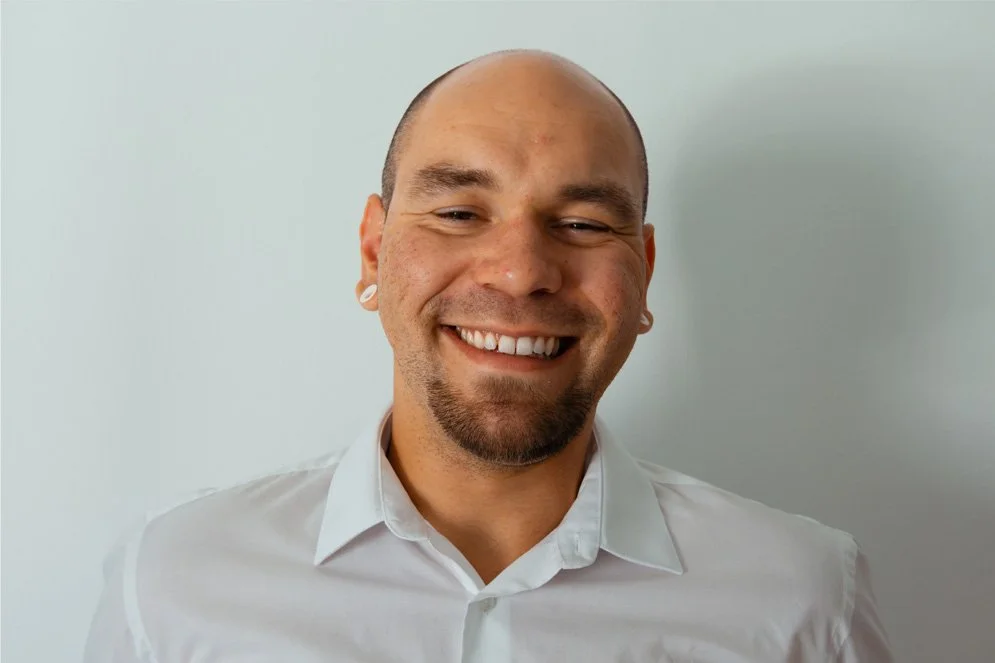 Smiling man with a goatee and earrings wearing a white shirt against a plain light background.