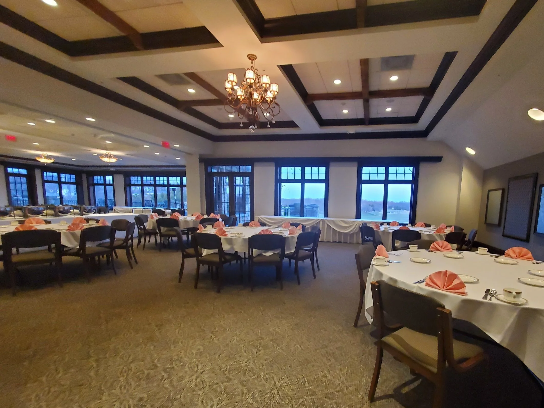 Elegant banquet hall with round tables set with white tablecloths, neatly folded pink napkins, and arranged silverware, featuring large windows and ornate chandeliers.