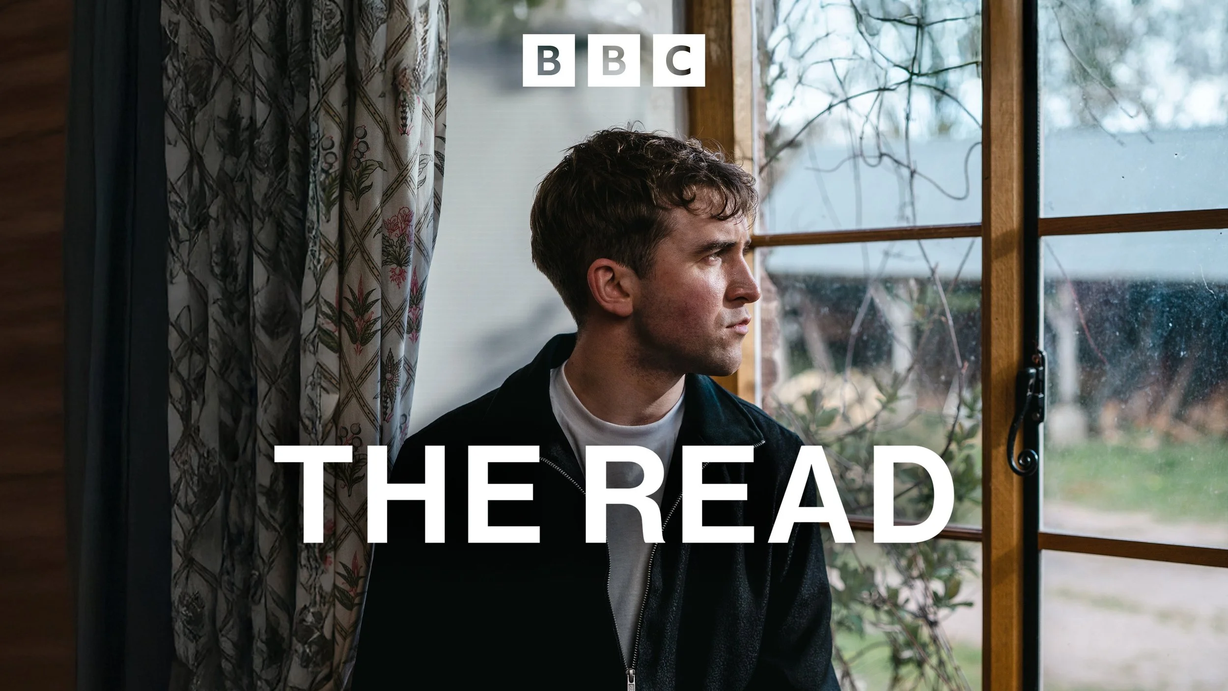 A young man with short, dark hair and a serious expression looking out a window with wooden framing, outside trees, and cloudy sky. Curtain with a floral pattern partially drawn to the side. Text overlay says "BBC THE READ."