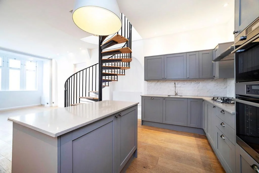 Modern kitchen with gray cabinets, marble countertop, spiral staircase, hanging lamp, and wood flooring.