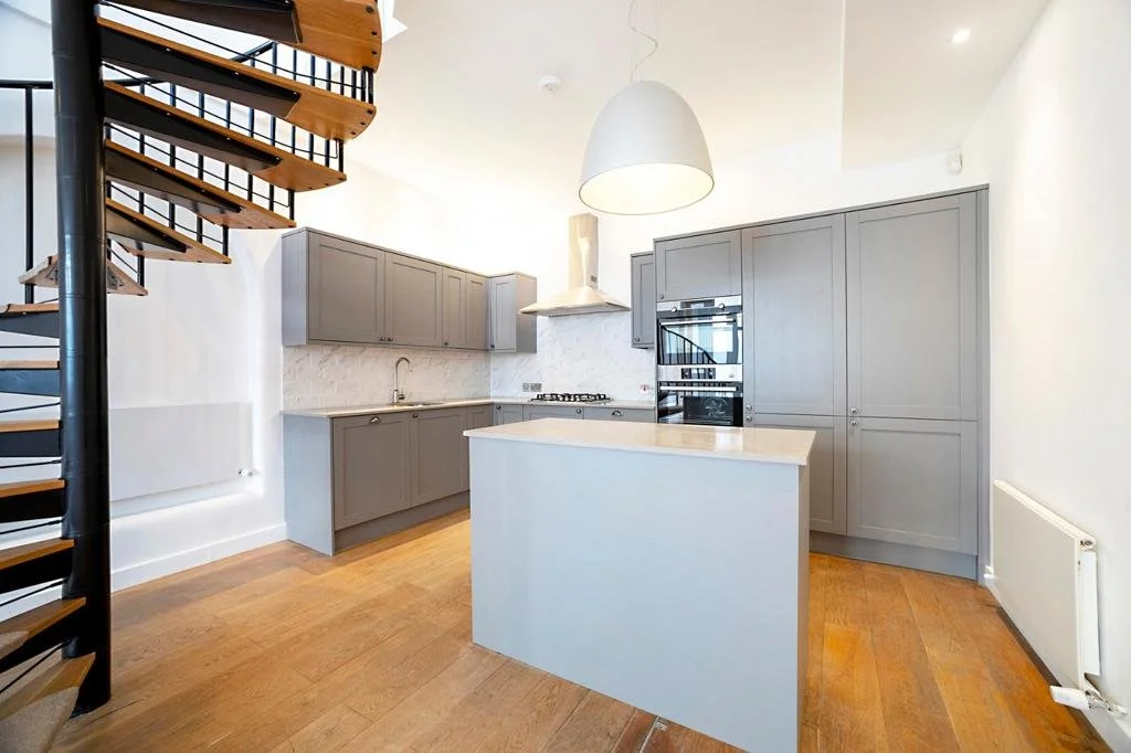 Modern kitchen with gray cabinets, island, wood floors, white walls, spiral staircase, and stainless steel appliances.