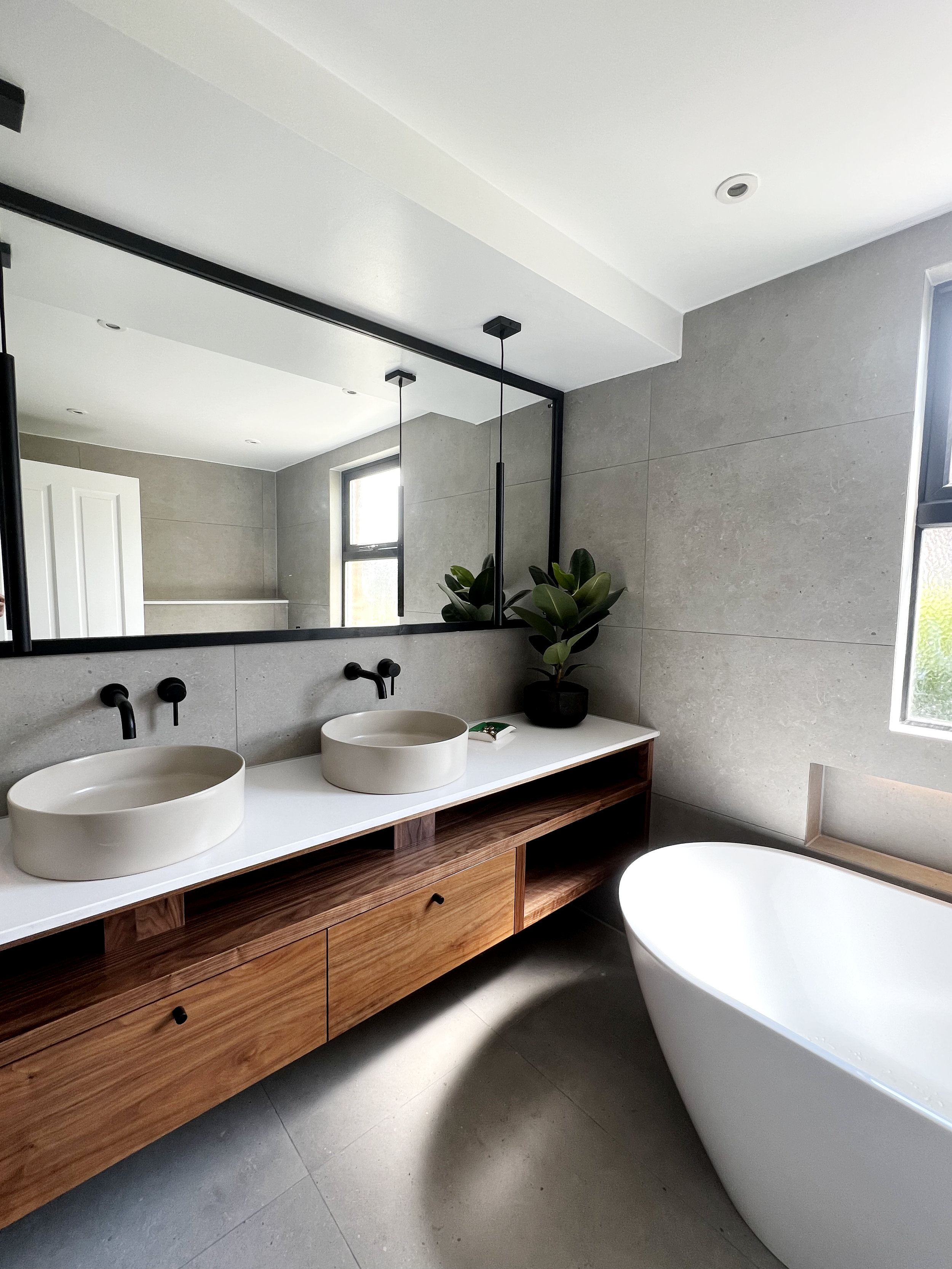 Modern bathroom with a large mirror, dual sinks, wooden vanity, freestanding bathtub, and potted plant.