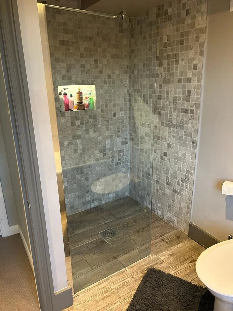 Modern bathroom with a walk-in shower featuring beige mosaic tiles, a glass partition, and a recessed shelf with various toiletries.