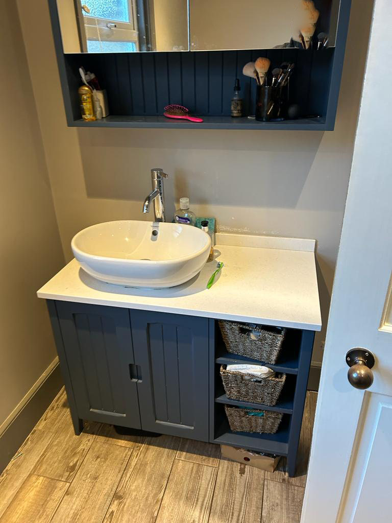 Bathroom with white sink, modern faucet, blue cabinet, wicker baskets, and toiletries. Shelves above hold brushes and personal care items.