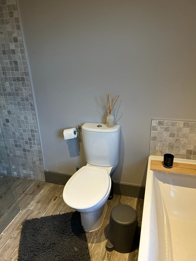 Bathroom interior featuring a toilet, toilet paper, a small black stool, a bath with a wooden ledge, a black candle, and a gray tiled shower area.
