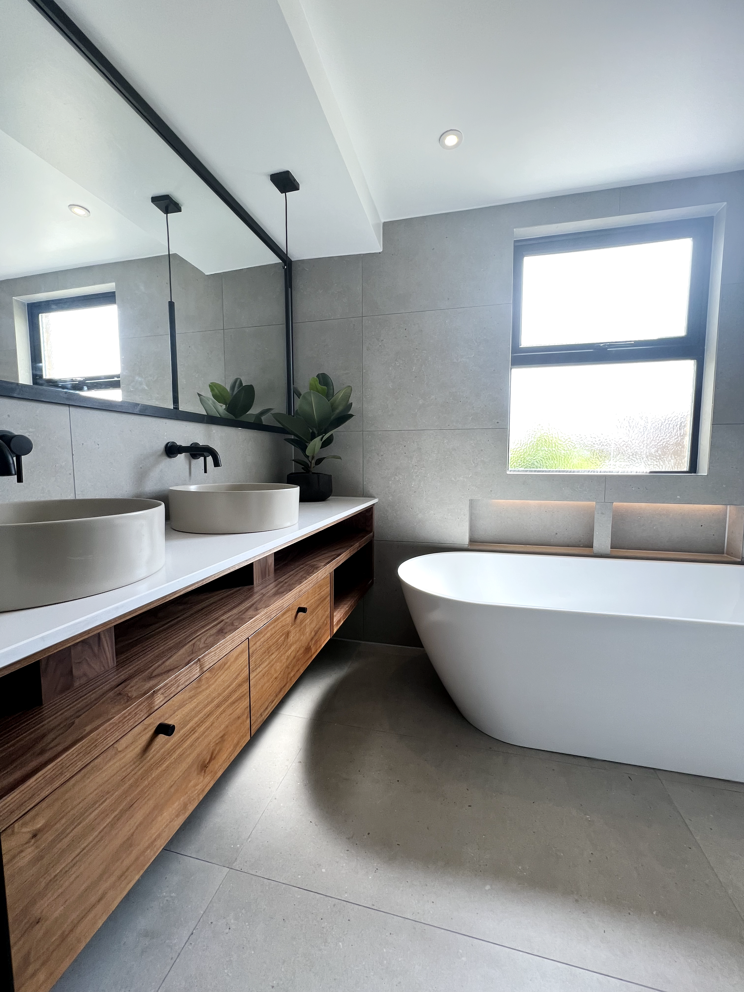 Modern bathroom interior with a freestanding bathtub, large mirror, double vessel sinks on a wooden vanity, tiled walls, and a window.