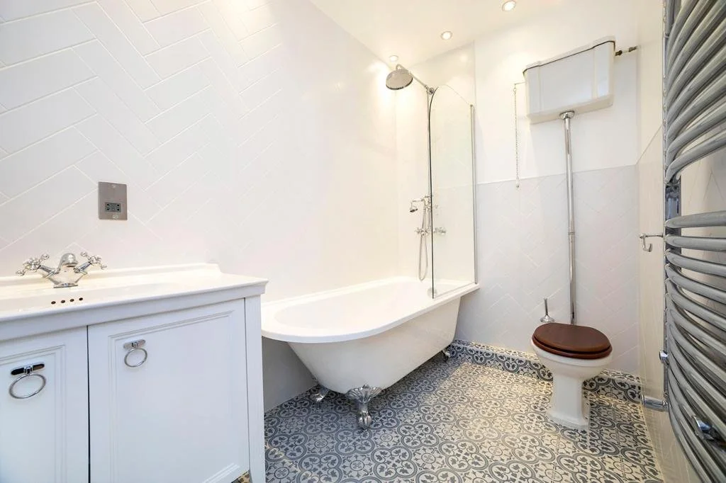 Bright bathroom with a freestanding bathtub, white sink, patterned floor tiles, wall-mounted toilet tank, and heated towel rail.