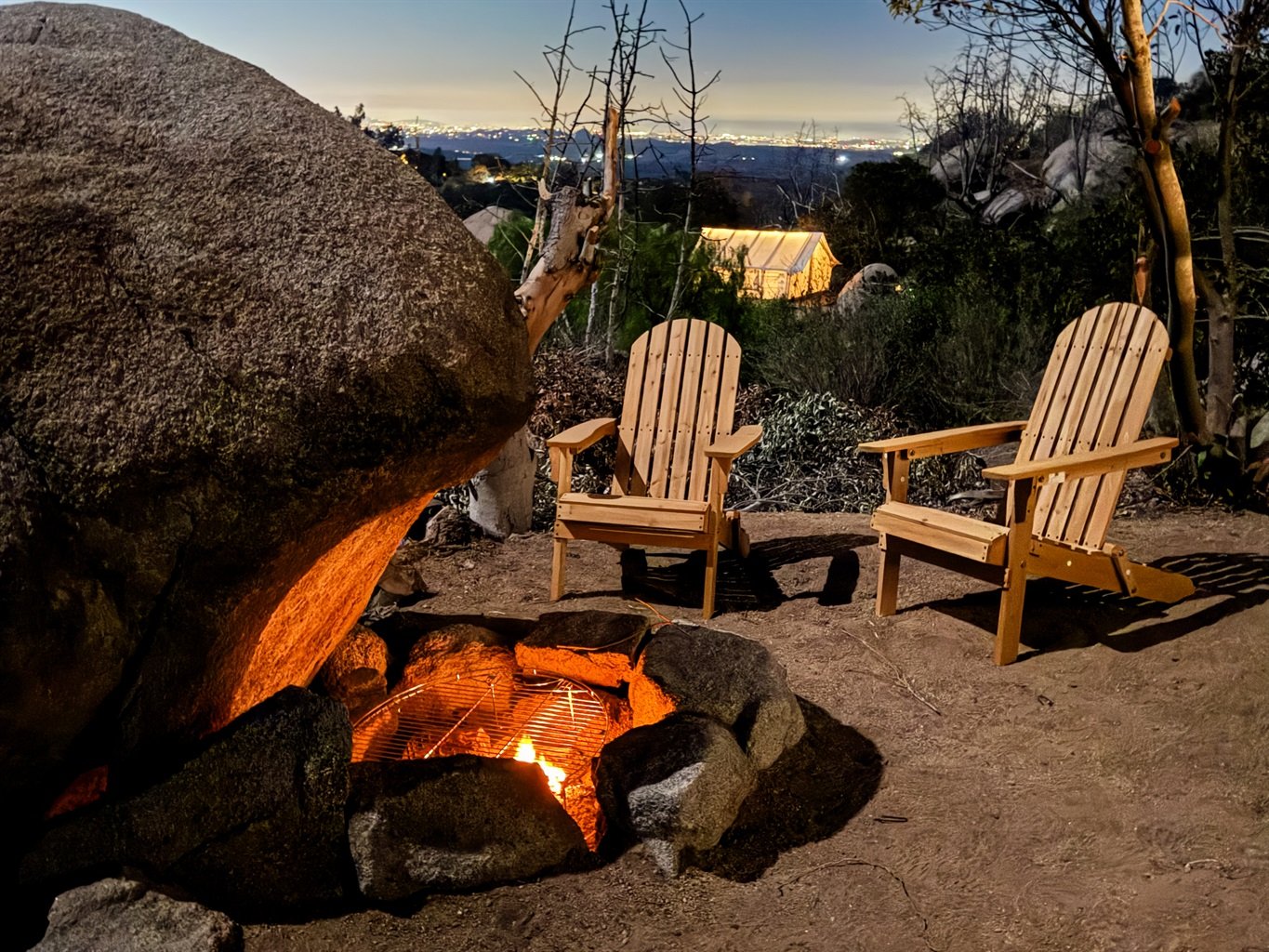Outdoor fire pit surrounded by large rocks with two wooden Adirondack chairs, nighttime setting, trees and distant building visible in the background.