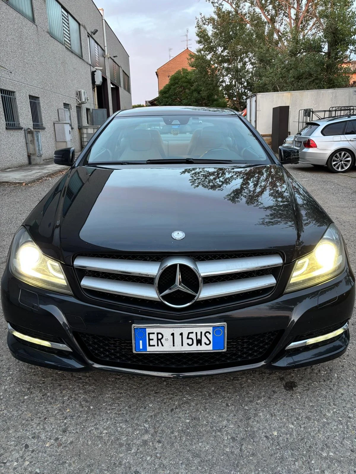 Auto Mercedes-Benz nero con fari accesi, vista frontale, con targa italiana ER 115WS, parcheggiata in un'area urbana con altri veicoli e edifici sullo sfondo.