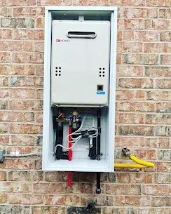 Outdoor wall-mounted tankless water heater with exposed pipes, mounted on brick wall.