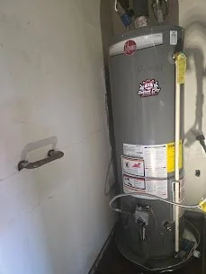 A water heater installed next to a light-colored wall with a towel bar and some wiring or pipes nearby.