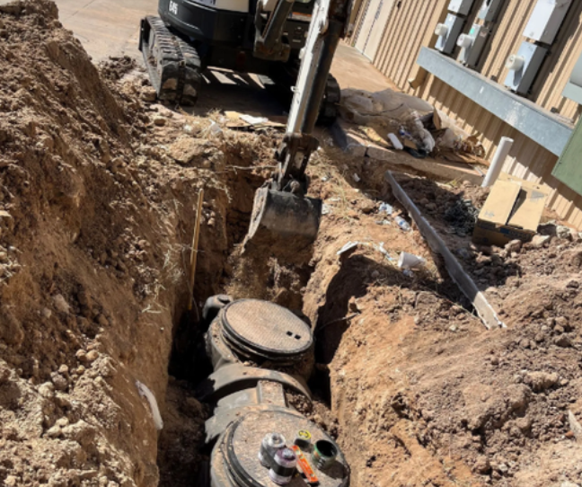 Excavator digging a trench with visible pipes and covers, surrounded by dirt and building materials.